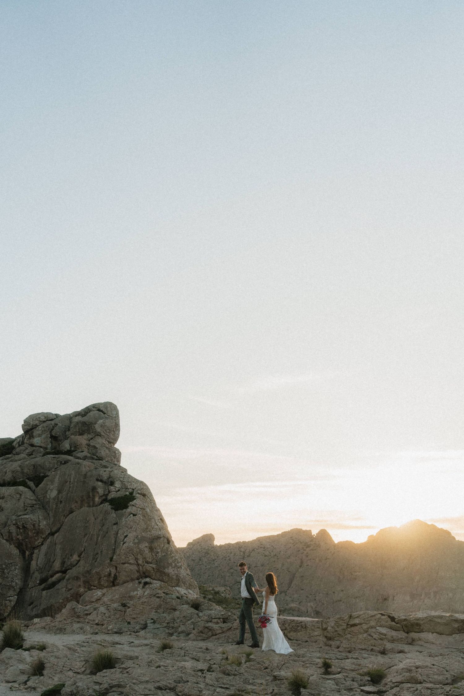 Wedding couple standing on rocky desert landscape at sunset, dramatic rock formations framing their silhouettes.