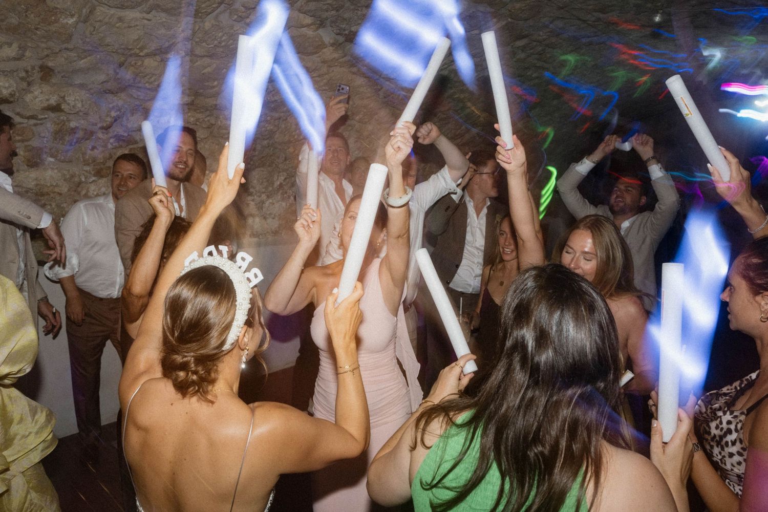 Excited wedding guests dancing and waving glowing LED foam sticks at a lively reception party.