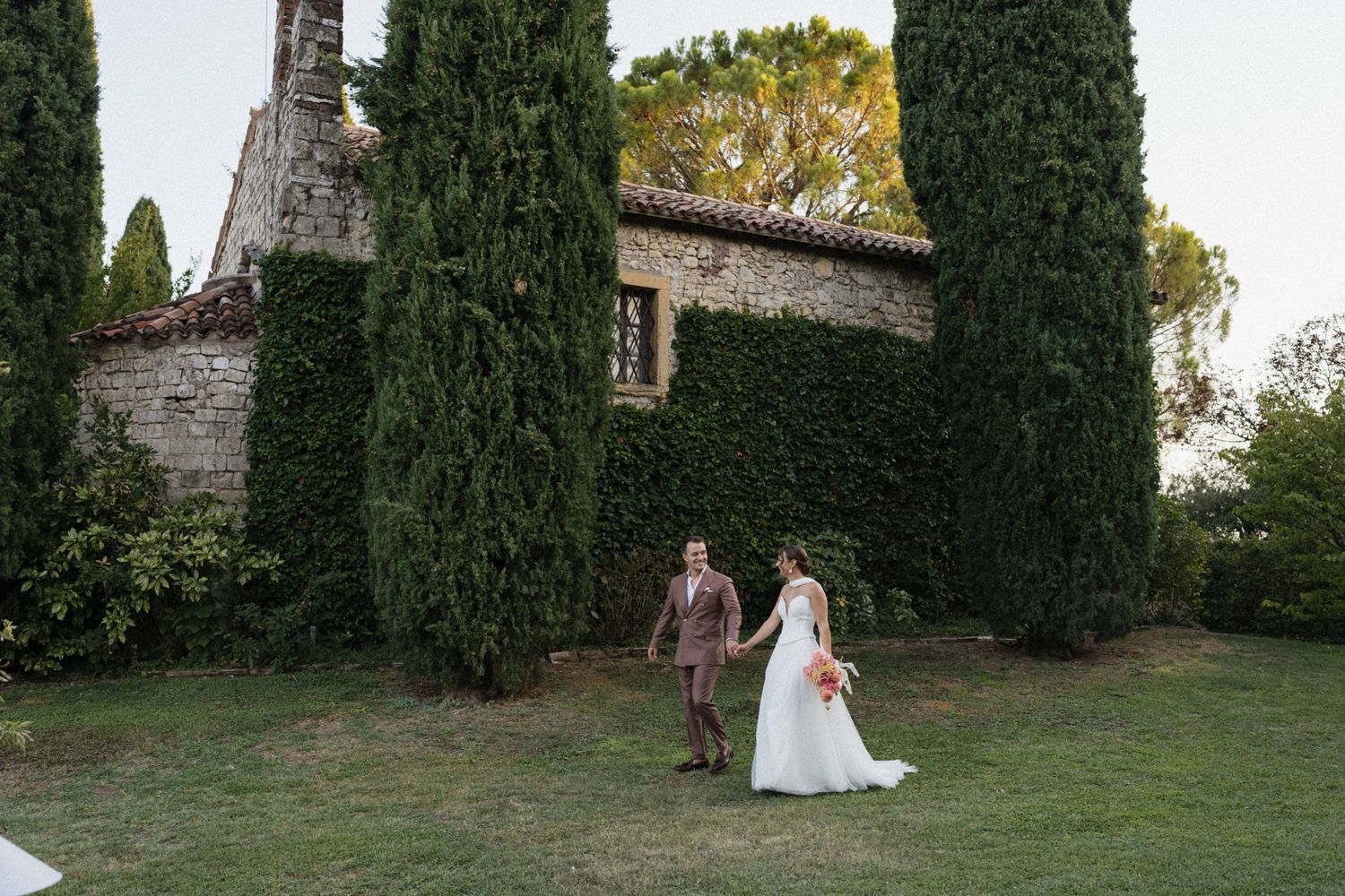 Bride and groom walk hand in hand on lush green lawn beside rustic stone chapel surrounded by tall cypress trees.