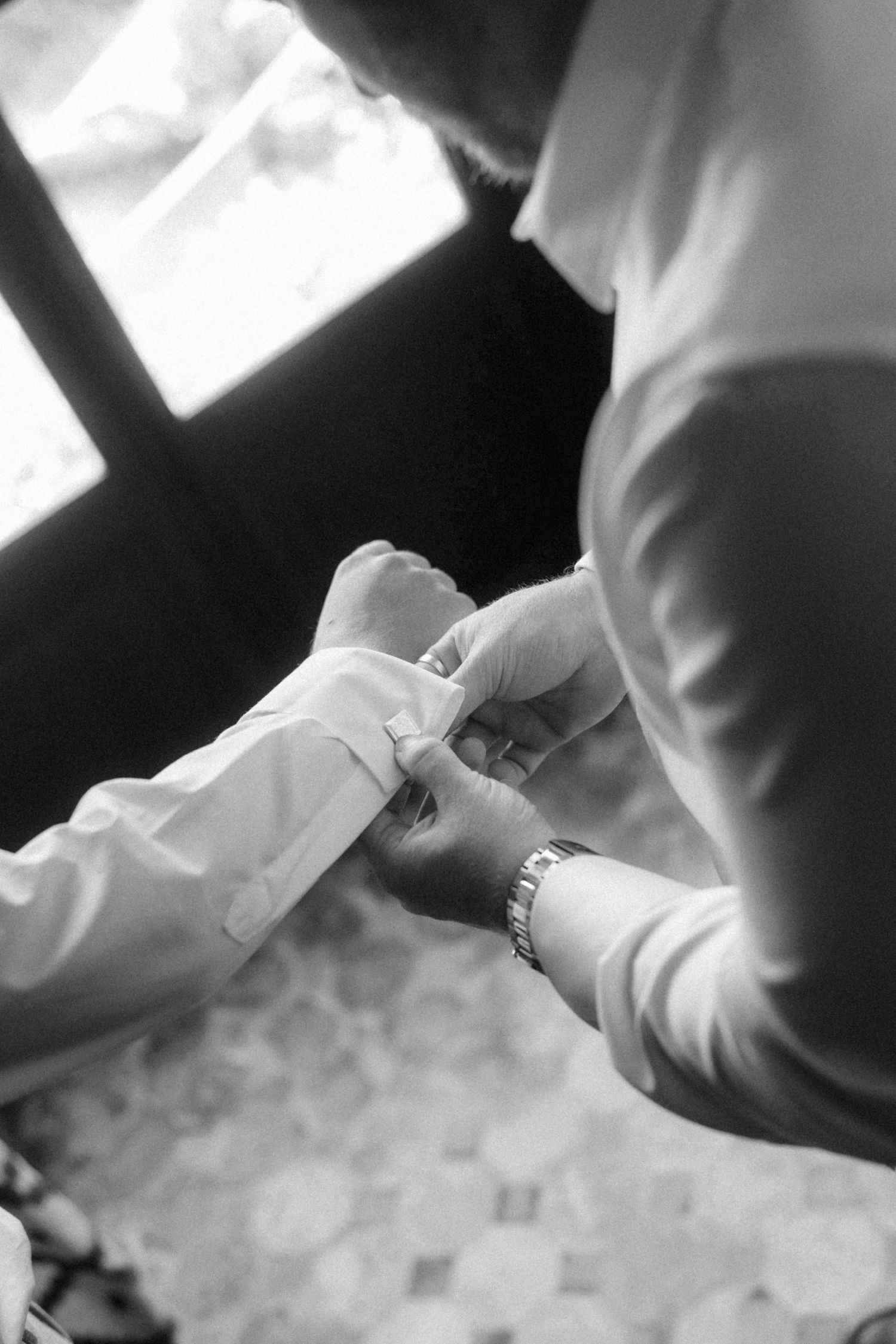 Black and white photo of a man fastening cufflinks on a white dress shirt before a wedding.
