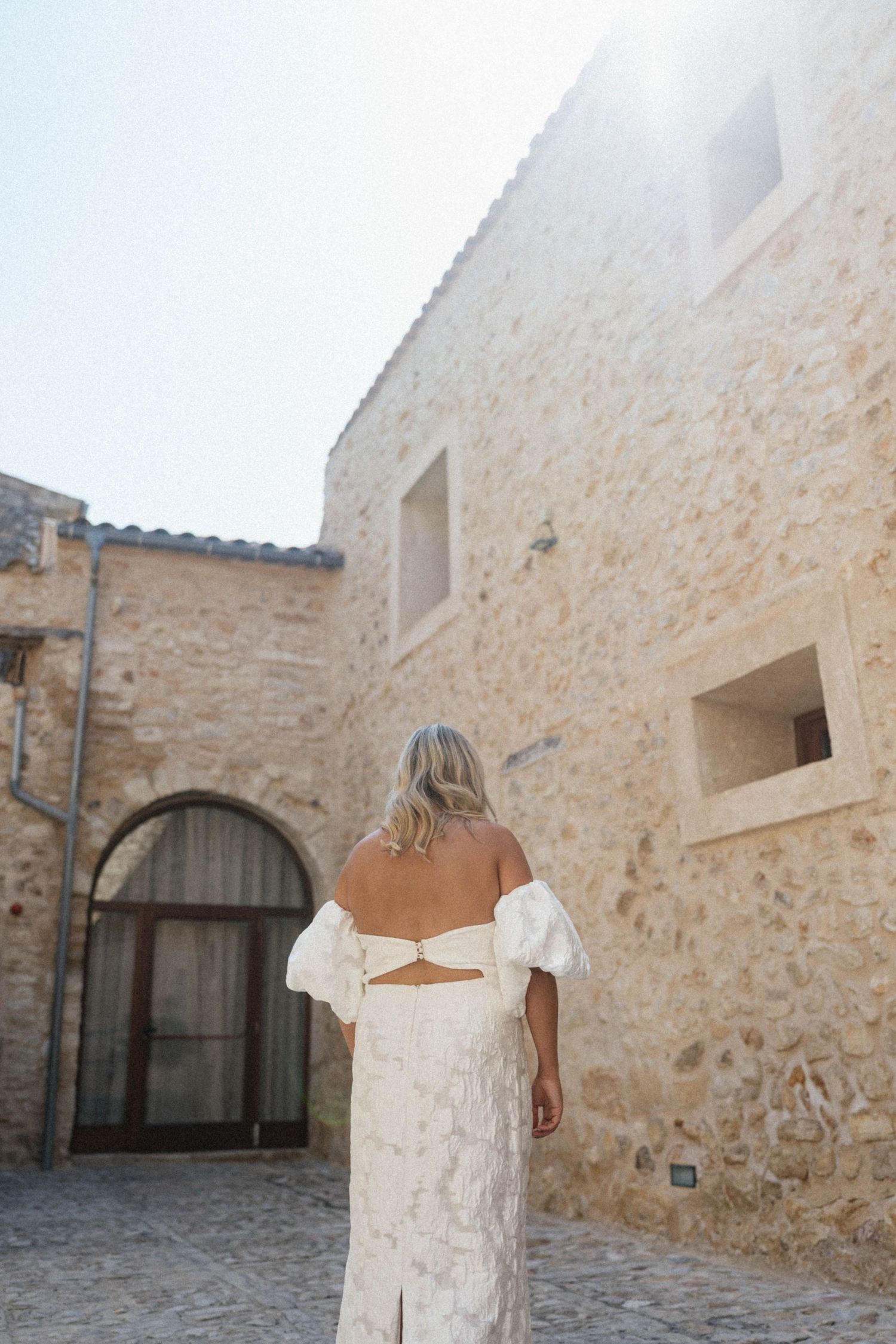 Bride in a backless white lace dress walking toward a sunlit rustic stone building.