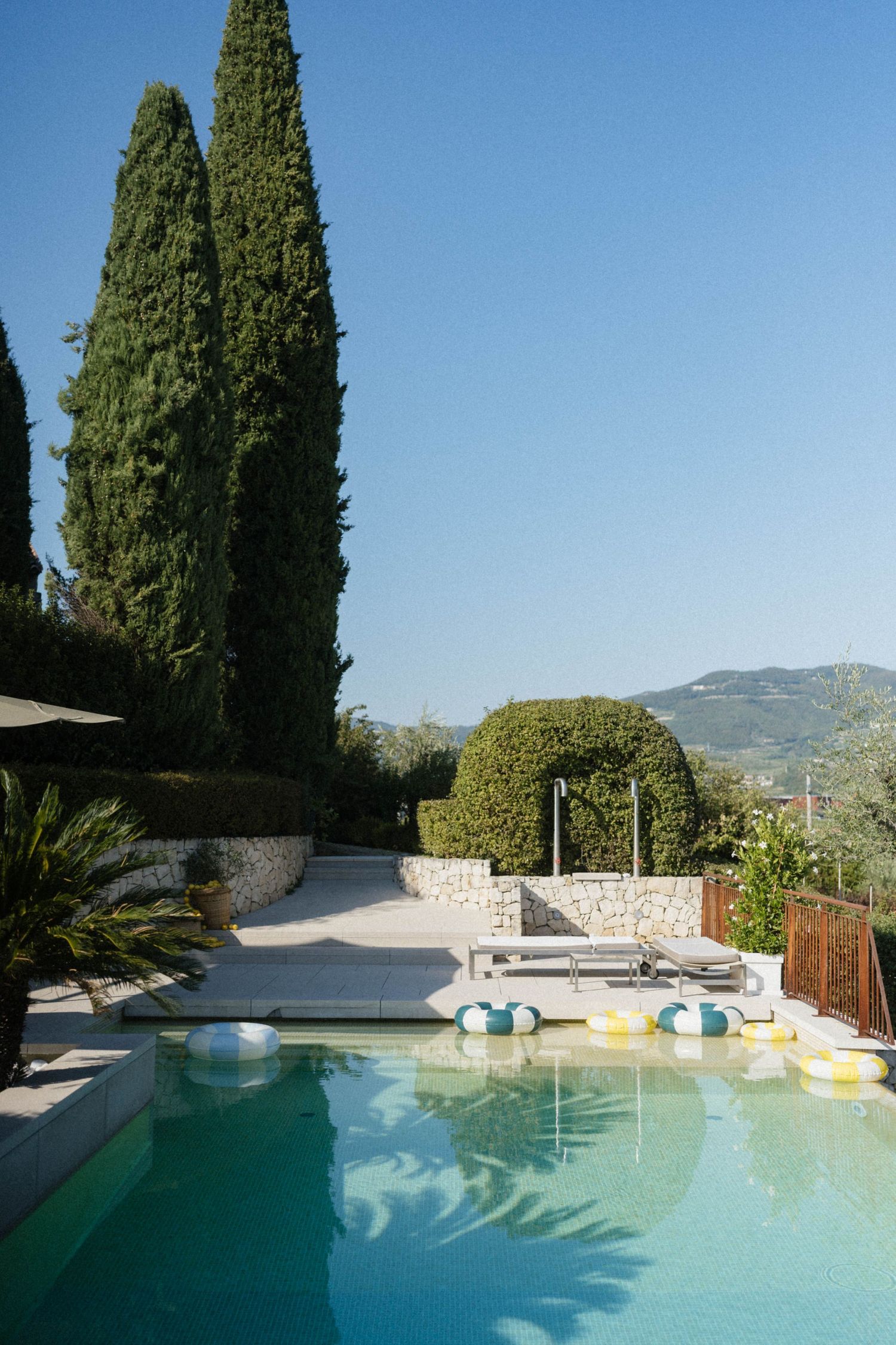 Luxury outdoor pool with cypress trees, lounge chairs, and mountain views under a clear blue sky.