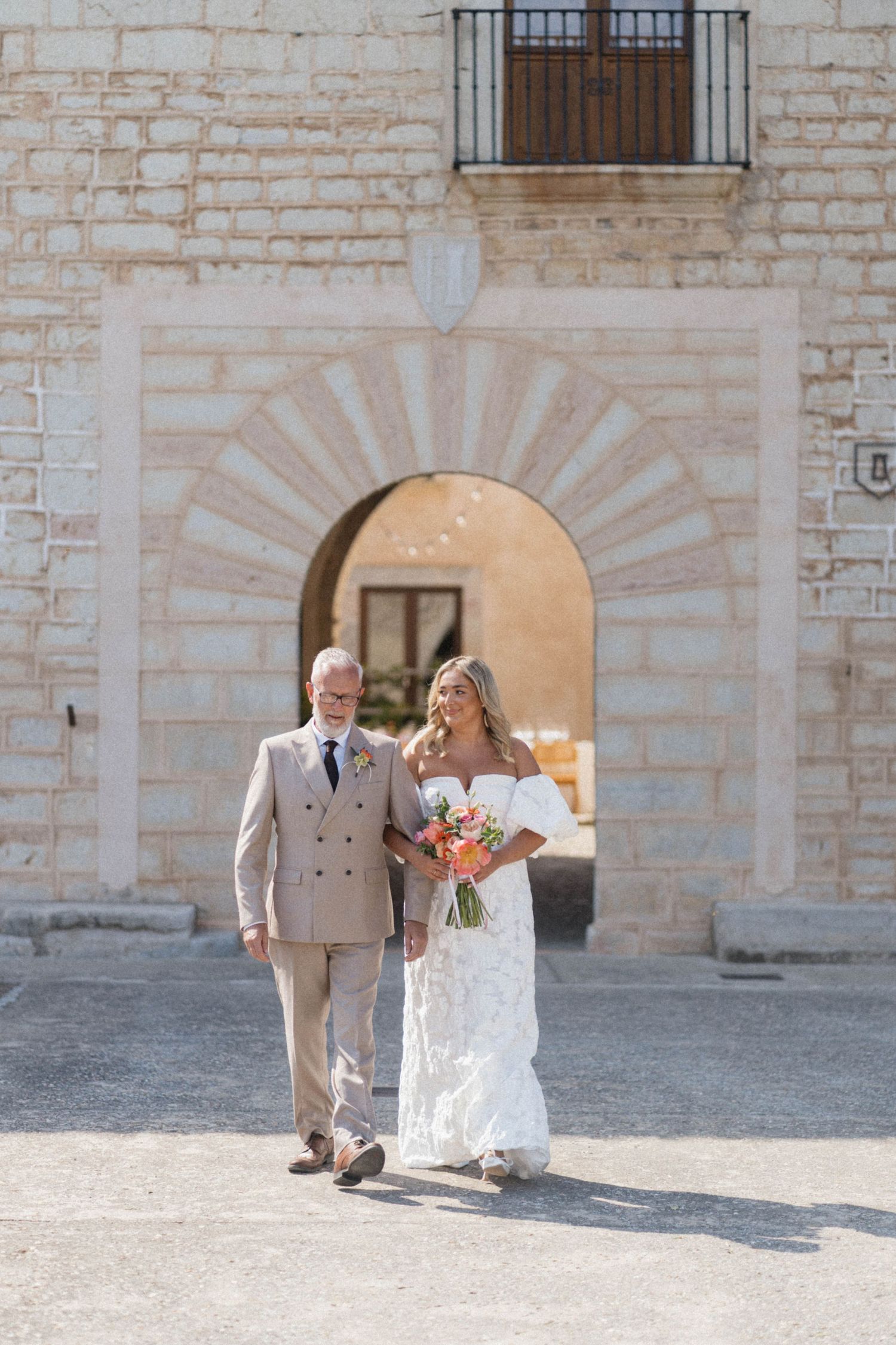 Bride in white lace gown and groom in tan suit walk together before a stone arch at an outdoor wedding venue.