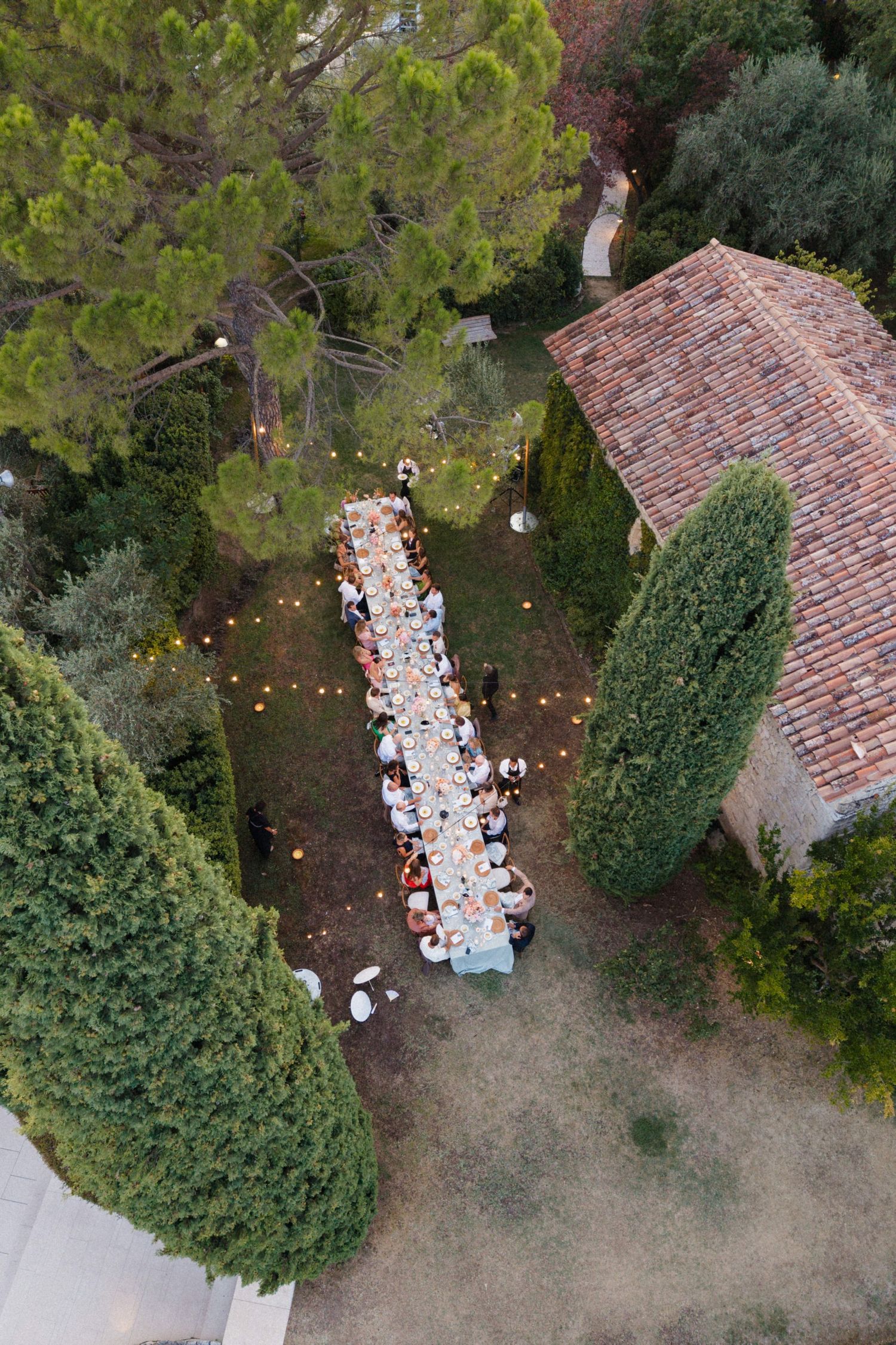 Aerial view of an elegant outdoor wedding reception with a long dining table surrounded by guests, set in a lush garden near a stone villa.