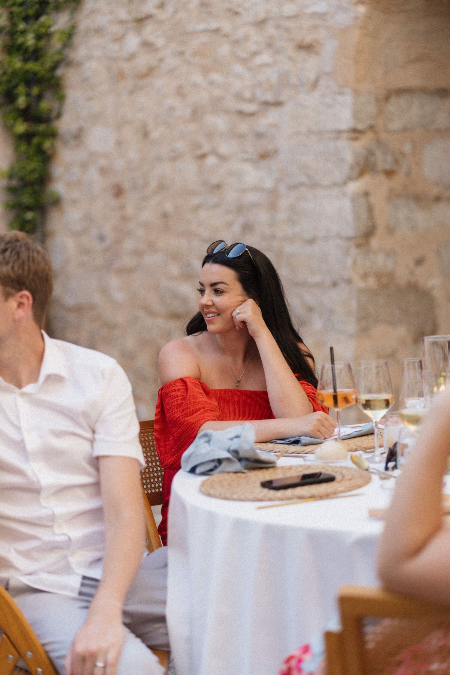 Woman in red off-shoulder dress smiling at outdoor wedding reception table with wine glasses.