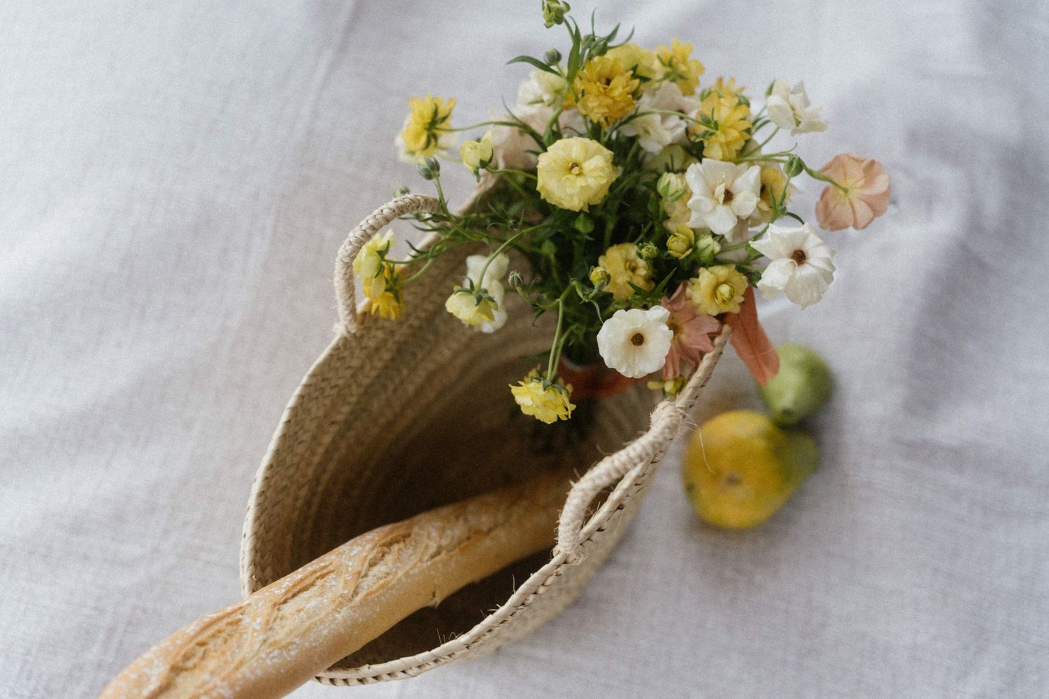 Straw tote bag with yellow and white wildflowers, a baguette, and green fruits on linen.