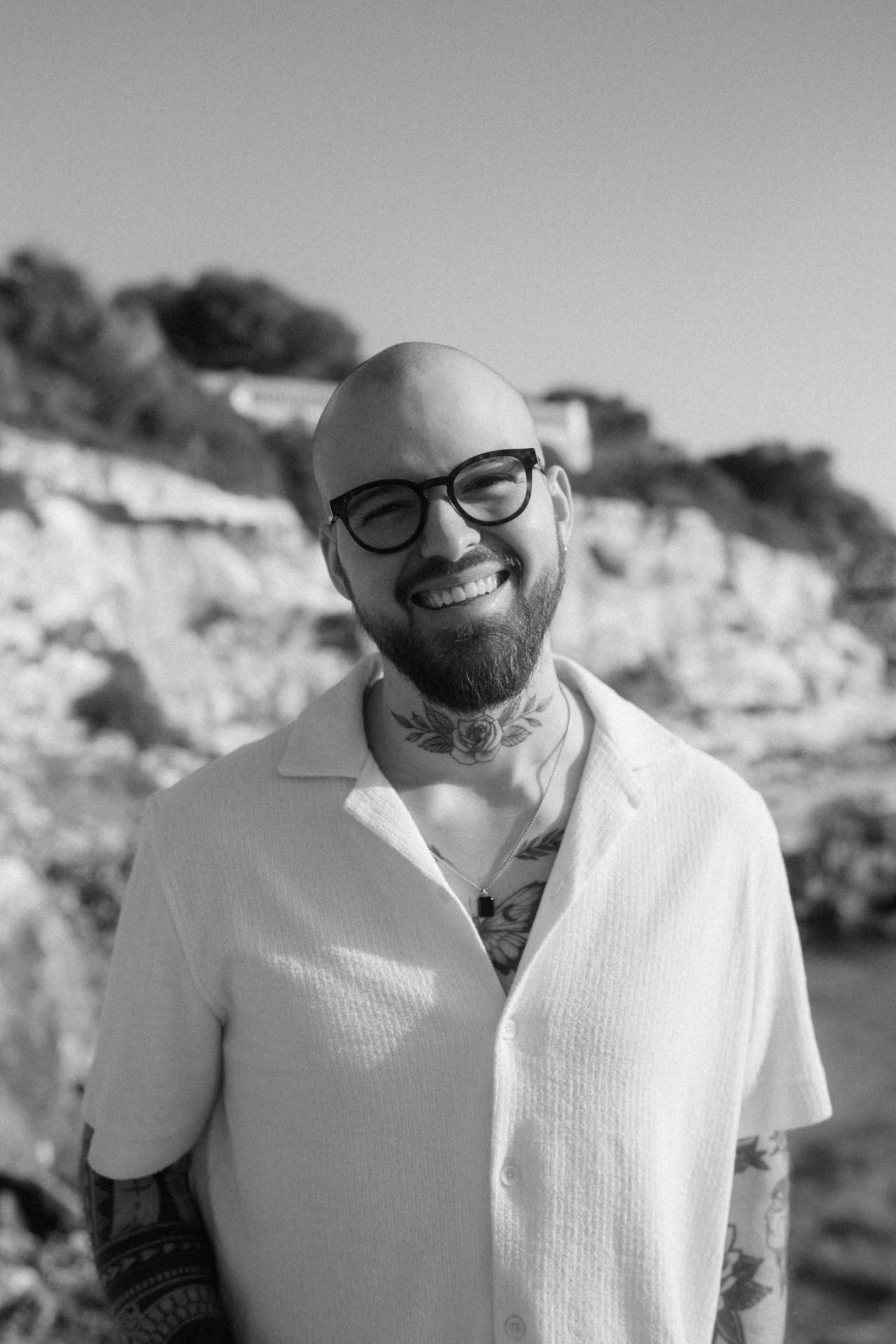 Smiling bald man with beard, glasses, and neck tattoos wearing a white shirt outdoors near rocky cliffs.