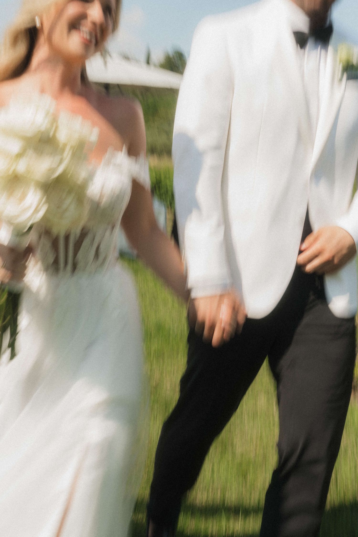 Bride in white off-shoulder gown holds bouquet while walking hand-in-hand with groom in white tuxedo outdoors.