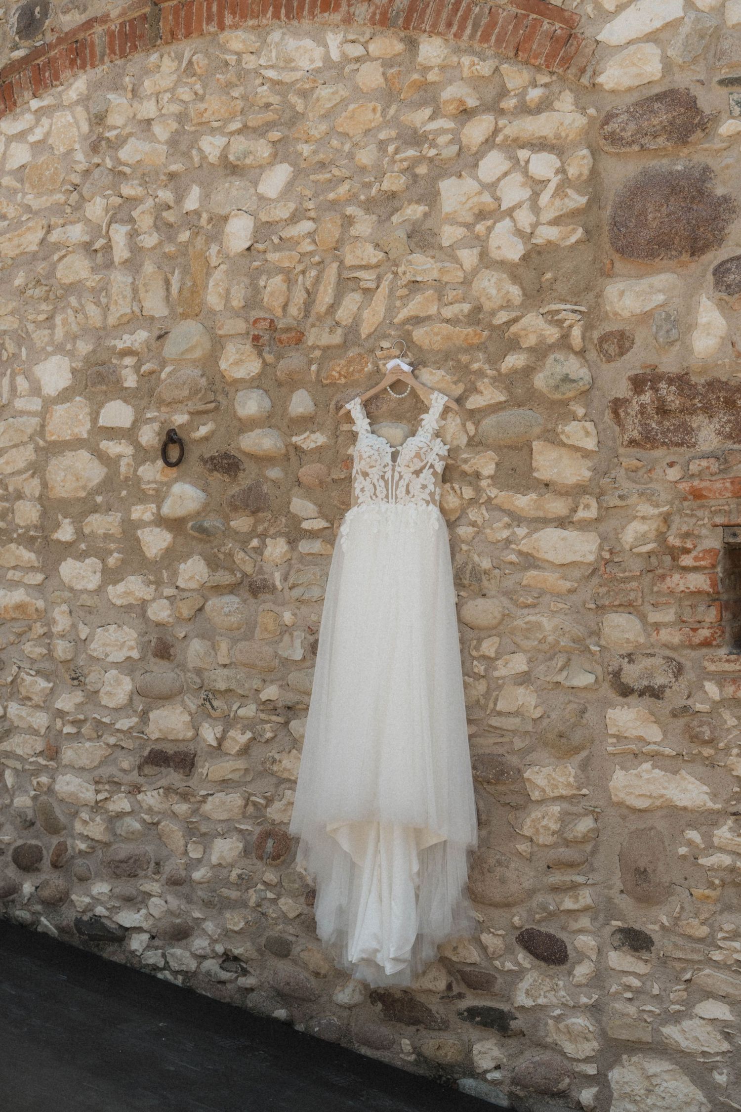 Elegant white lace wedding dress hanging on a rustic stone wall.