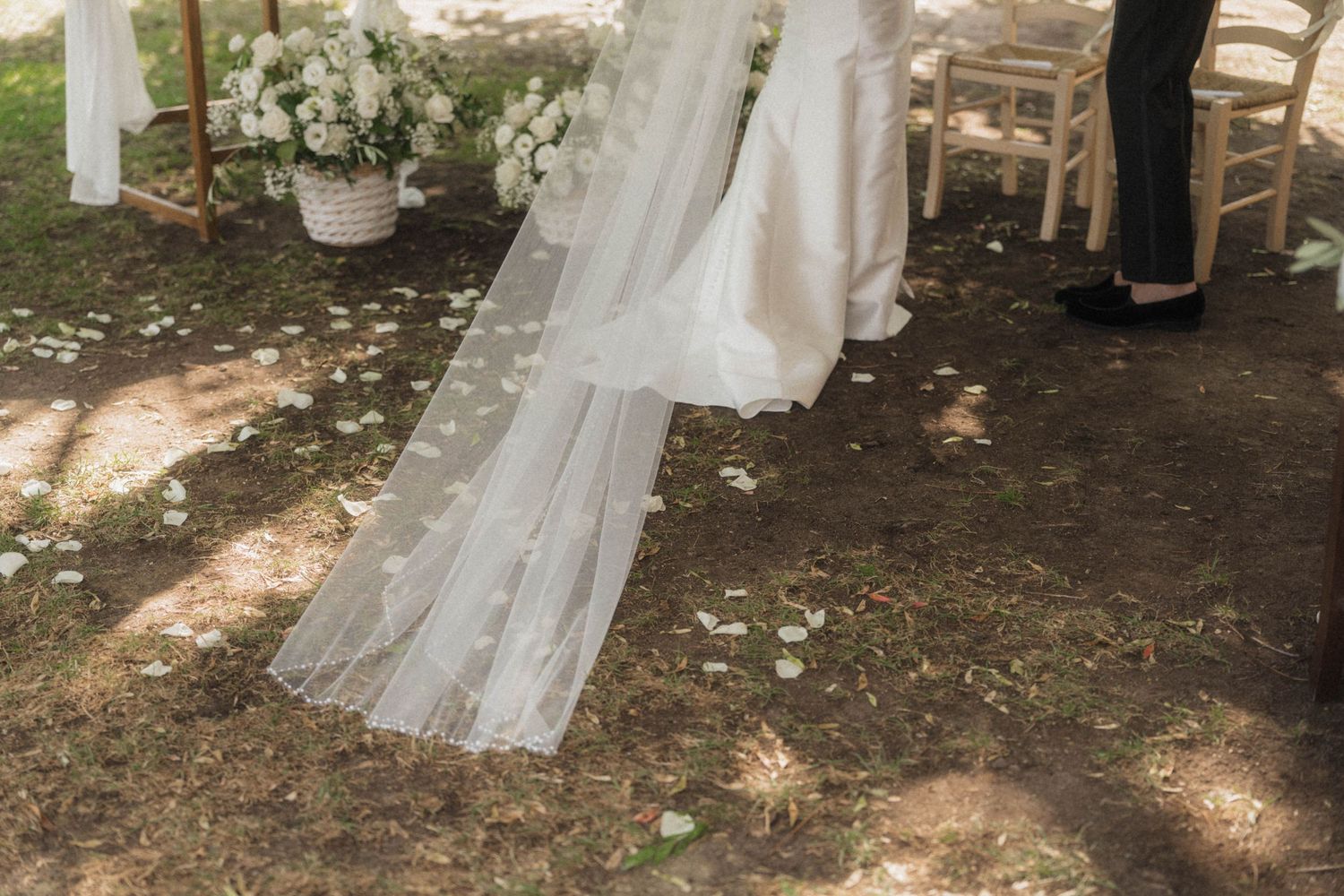 Bride in white gown with long lace veil trailing on ground at outdoor wedding ceremony with flower petals.