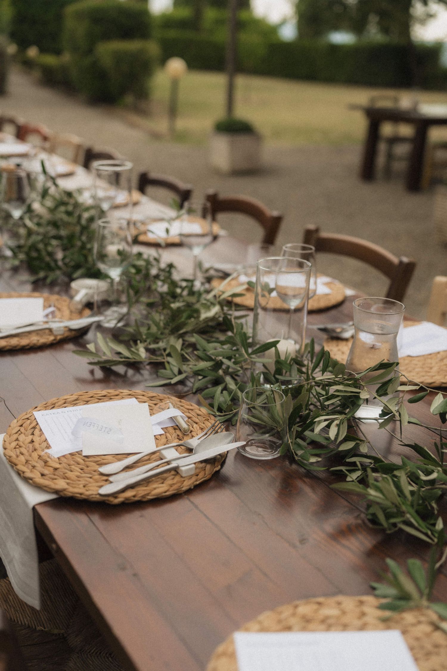 Elegant outdoor wedding reception table with woven placemats, greenery runner, and glassware.