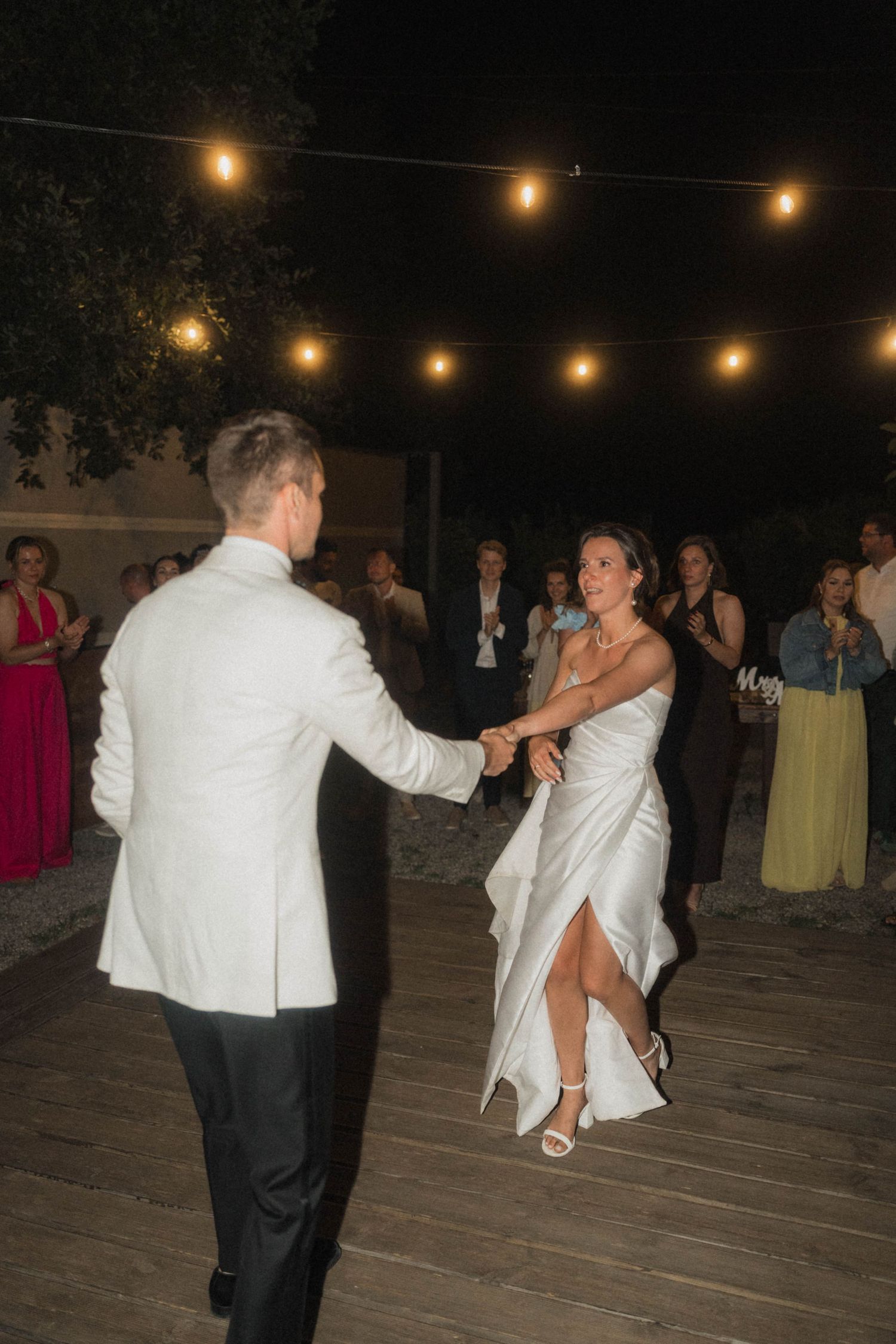 Bride and groom sharing first dance on outdoor wooden floor under warm string lights at night.