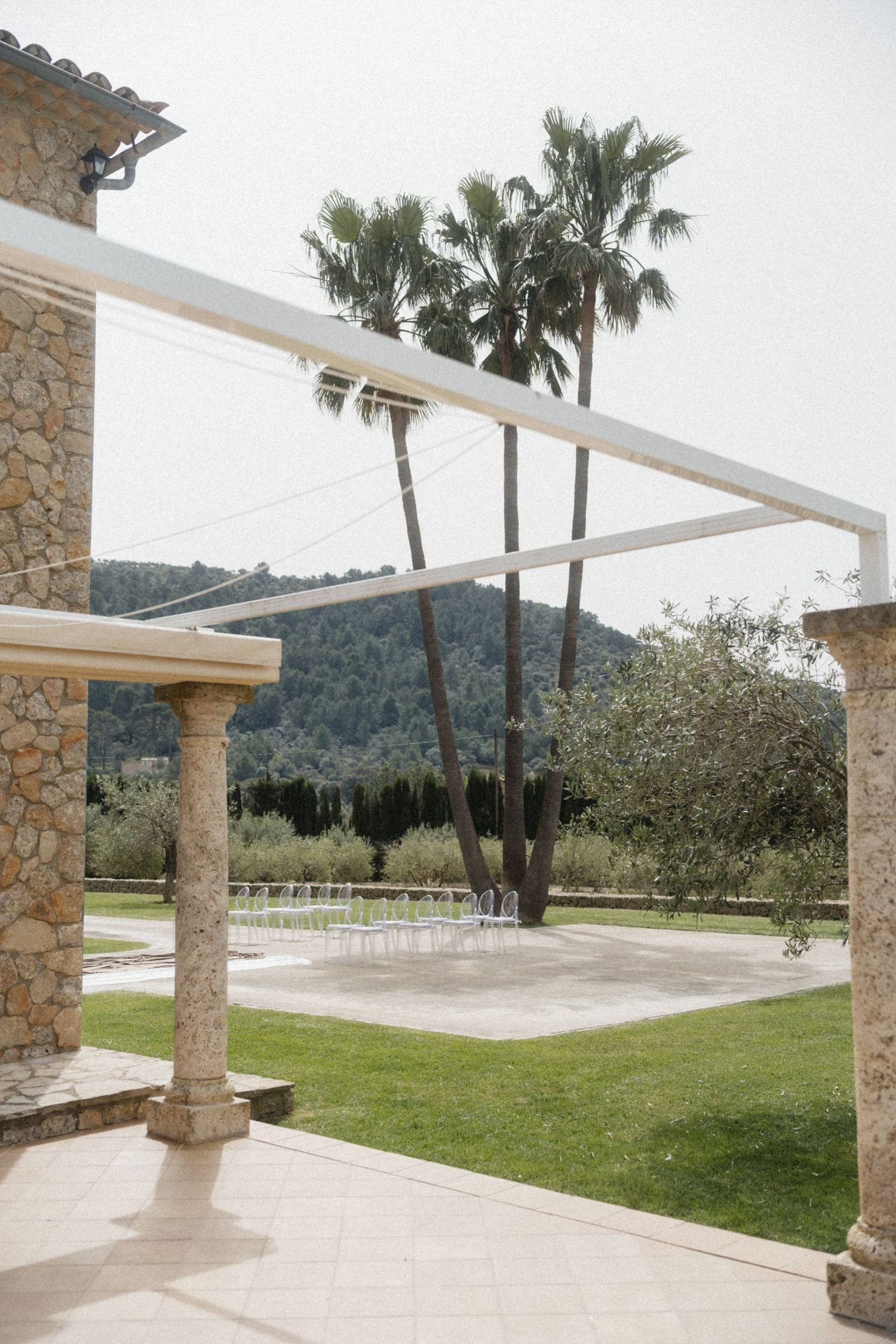 Stone villa pergola with palm trees and mountain views, elegant outdoor wedding ceremony setup on manicured grounds.