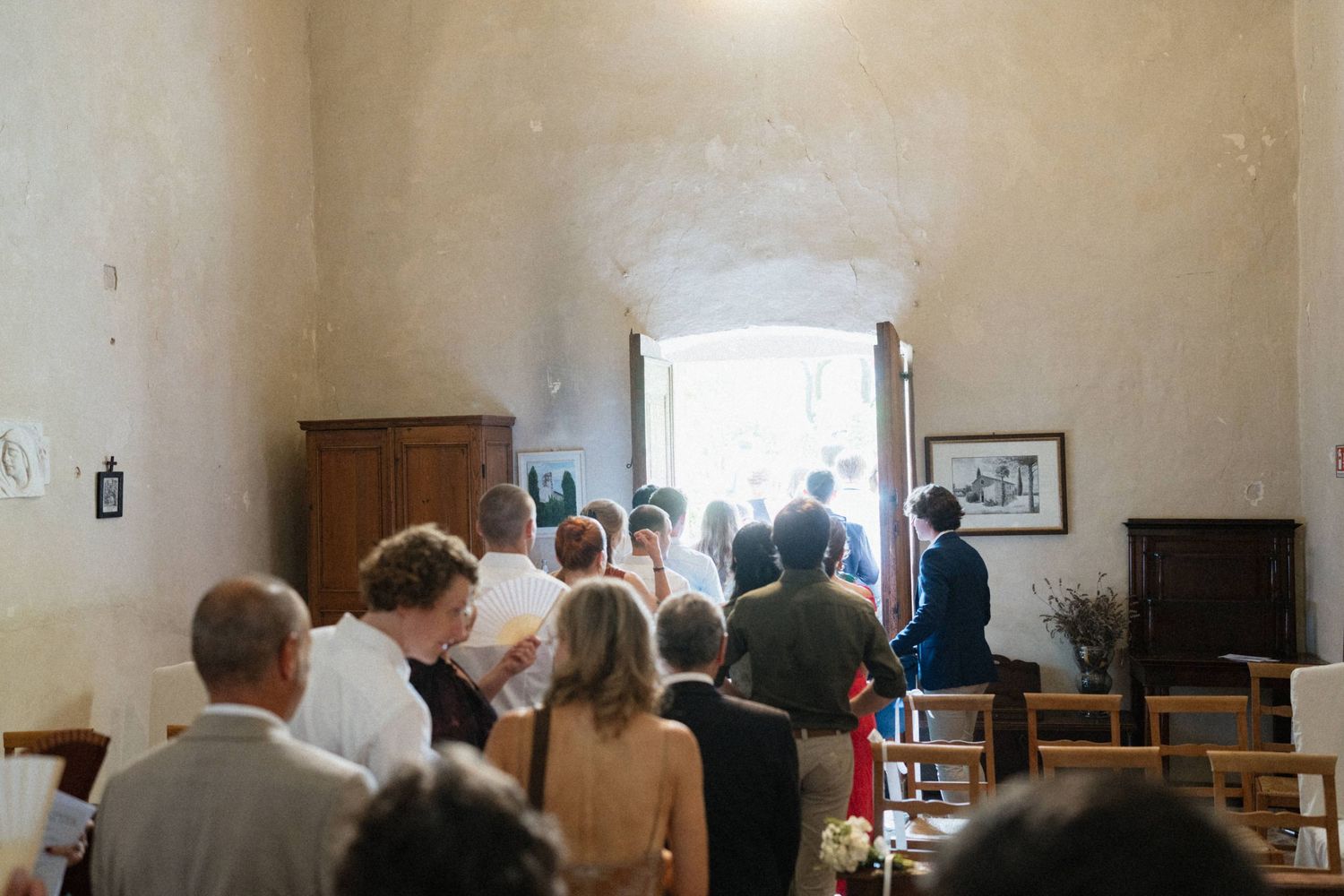 Wedding guests gathered in a rustic venue with stone walls, walking toward an open door filled with natural light.