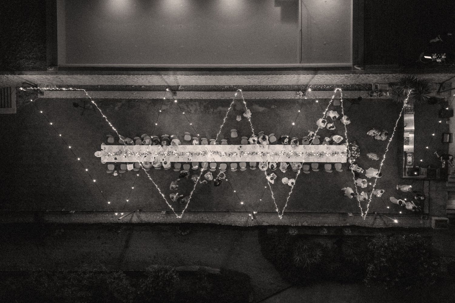 Black and white aerial photo of a wedding reception with zigzag string lights illuminating a long banquet table.