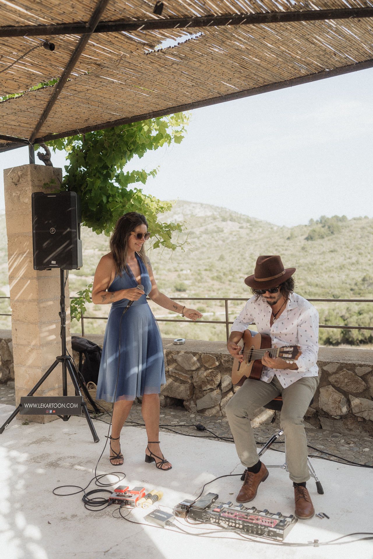 A woman in a blue dress and a guitarist in a brown hat perform outdoors under a thatched roof with scenic hills.