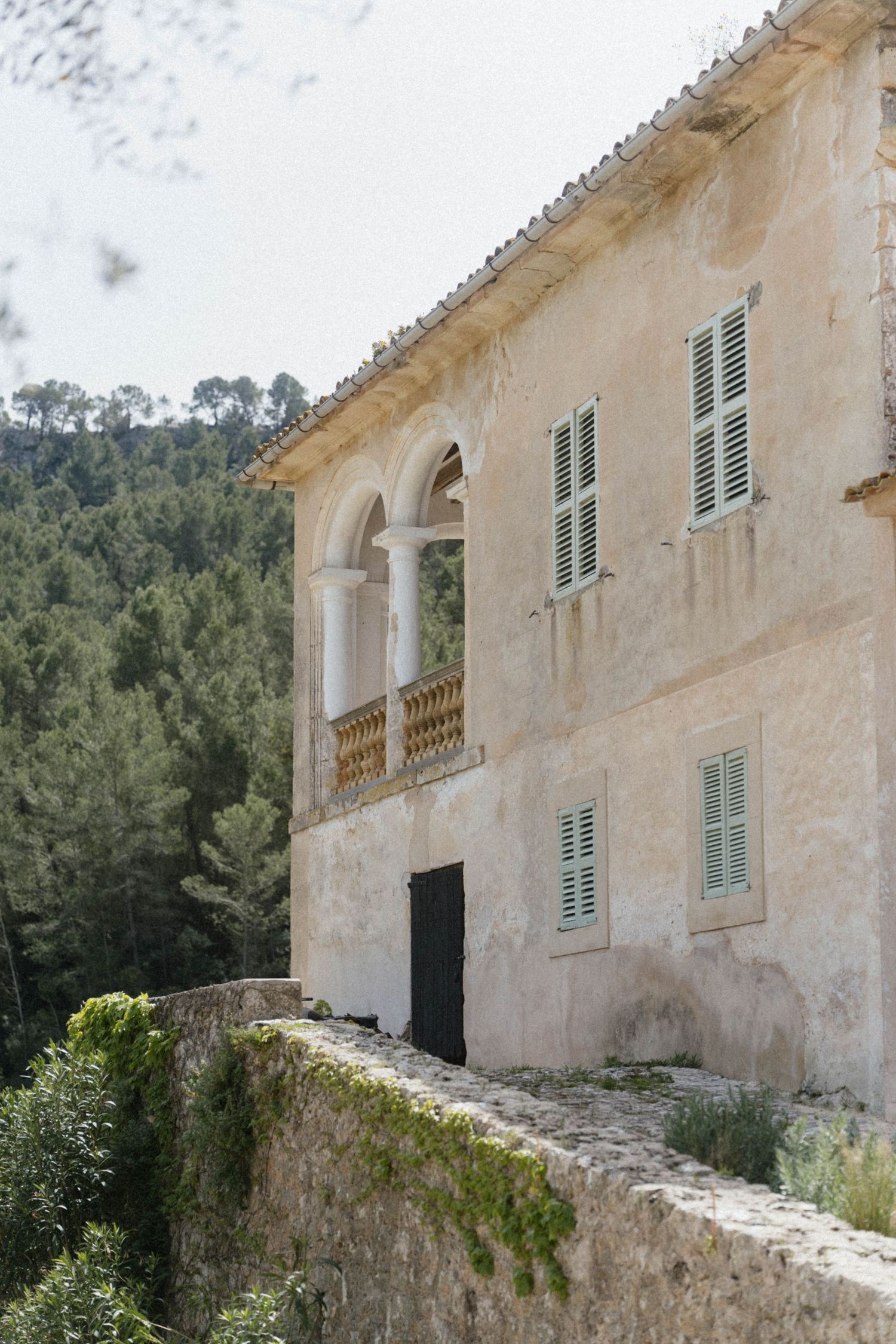 Rustic Mediterranean villa with weathered stone walls, arched balcony, and green shuttered windows surrounded by lush forest.