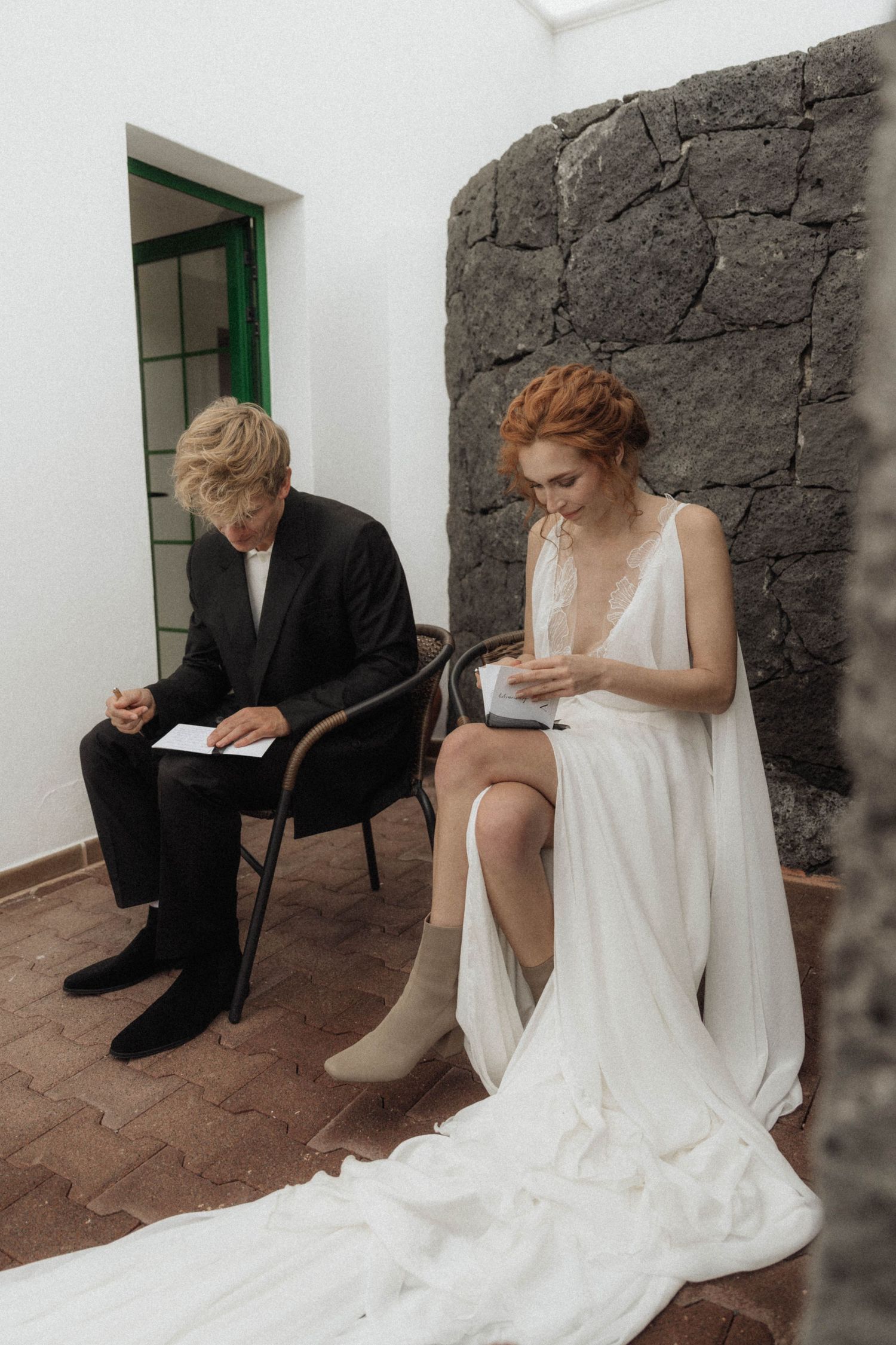 Bride and groom sitting on chairs reading vows before wedding ceremony indoors.