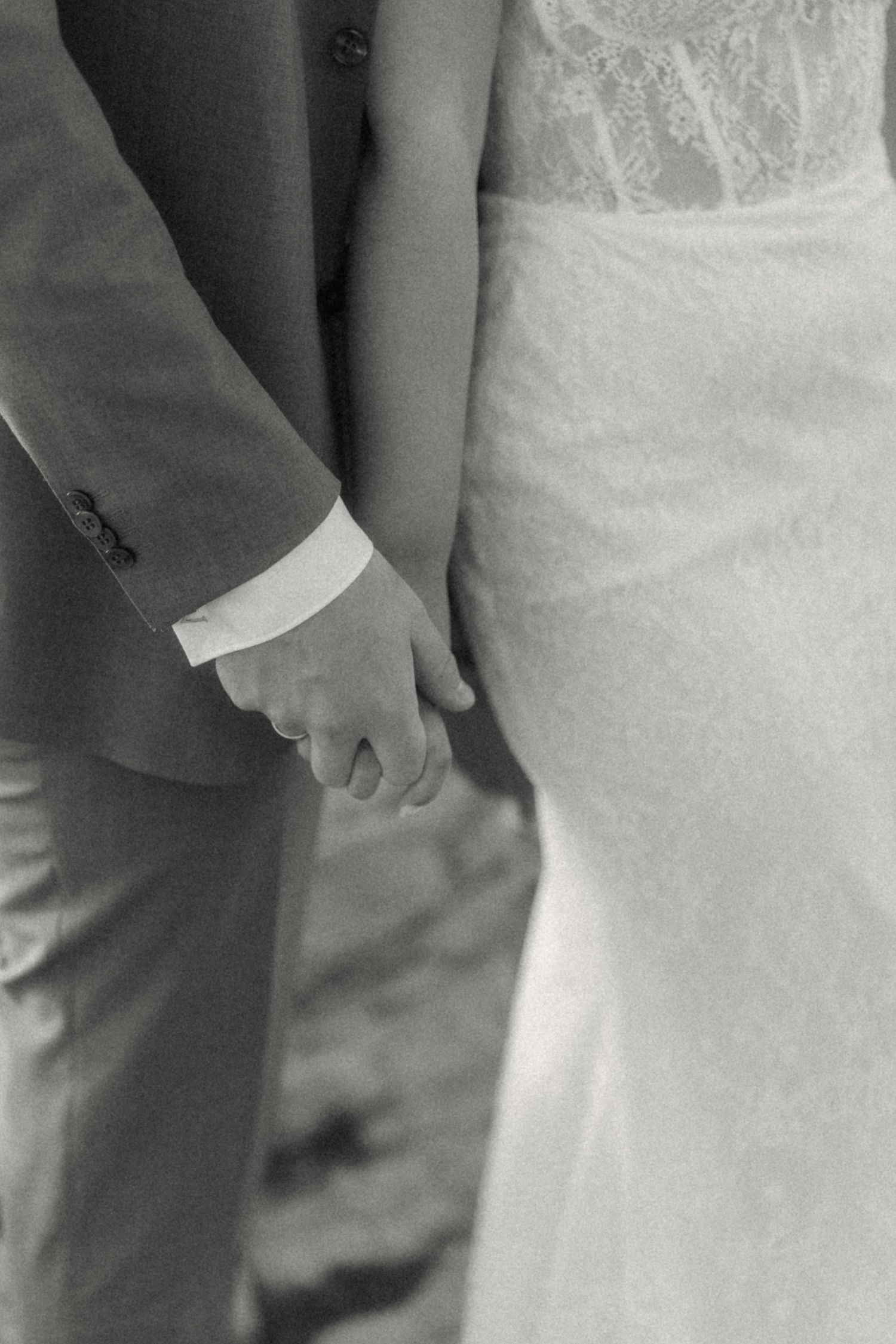 Close-up black and white photo of a bride and groom holding hands, showcasing lace wedding dress detail.