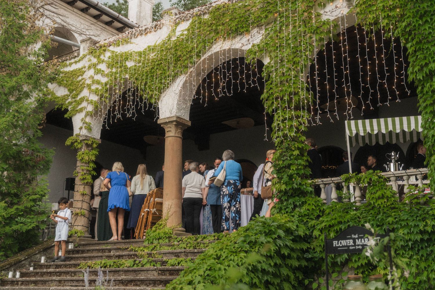 Wedding guests gather on ivy-covered stone terrace entrance of an elegant historic venue at golden hour.