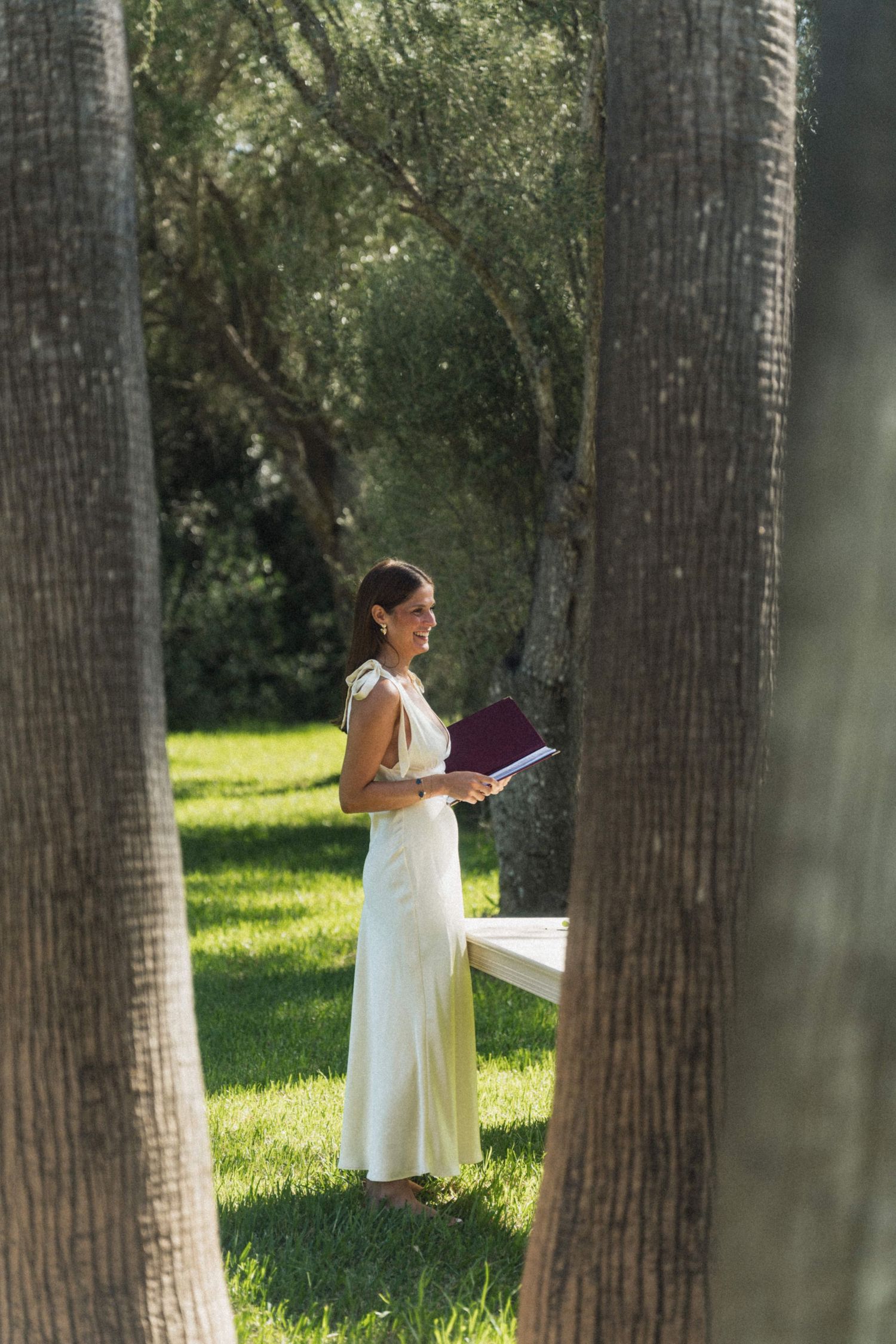 A woman in a white dress reads from a book while standing between palm trees at an outdoor wedding.