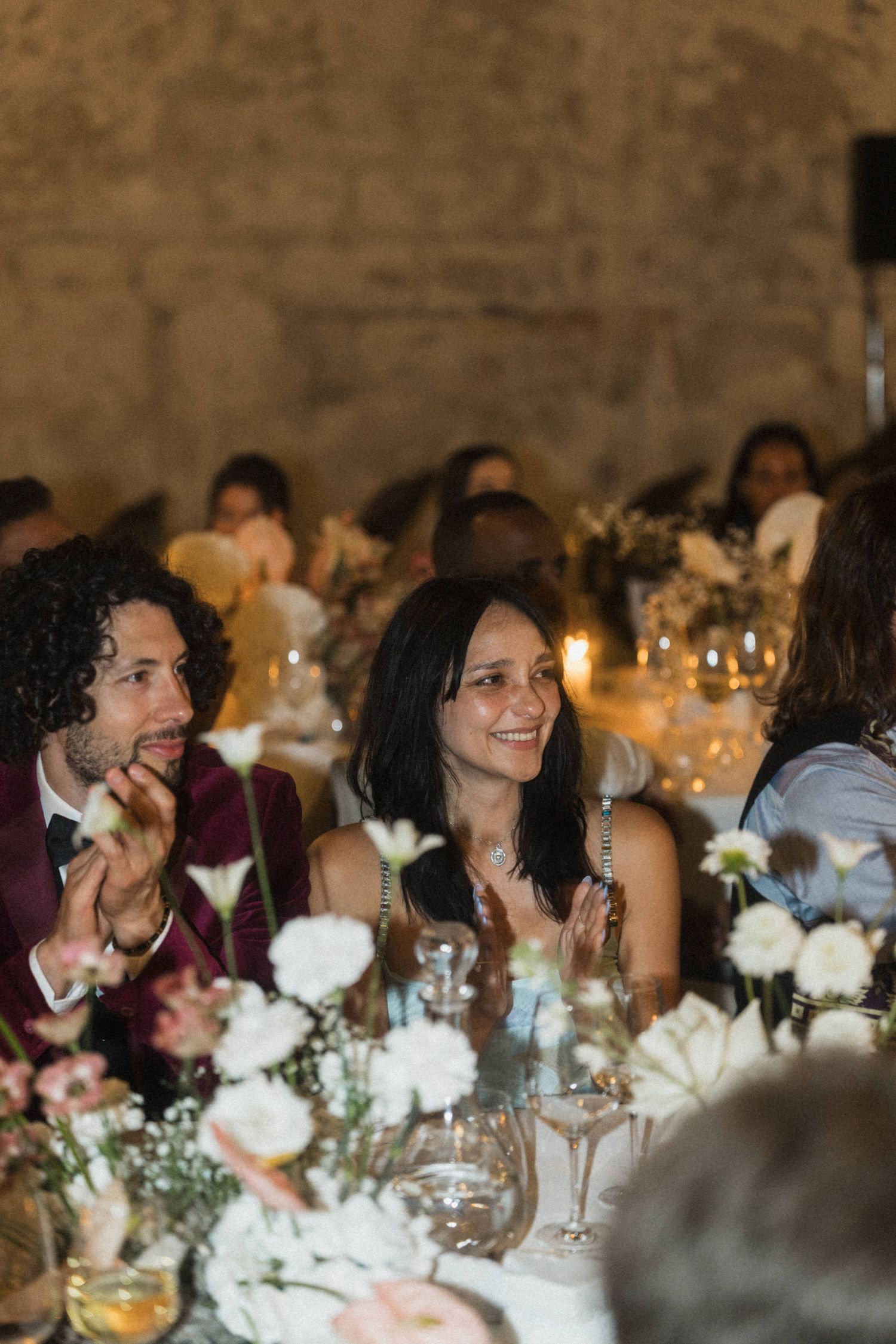 Smiling wedding guests clap at candlelit reception table adorned with white floral arrangements.