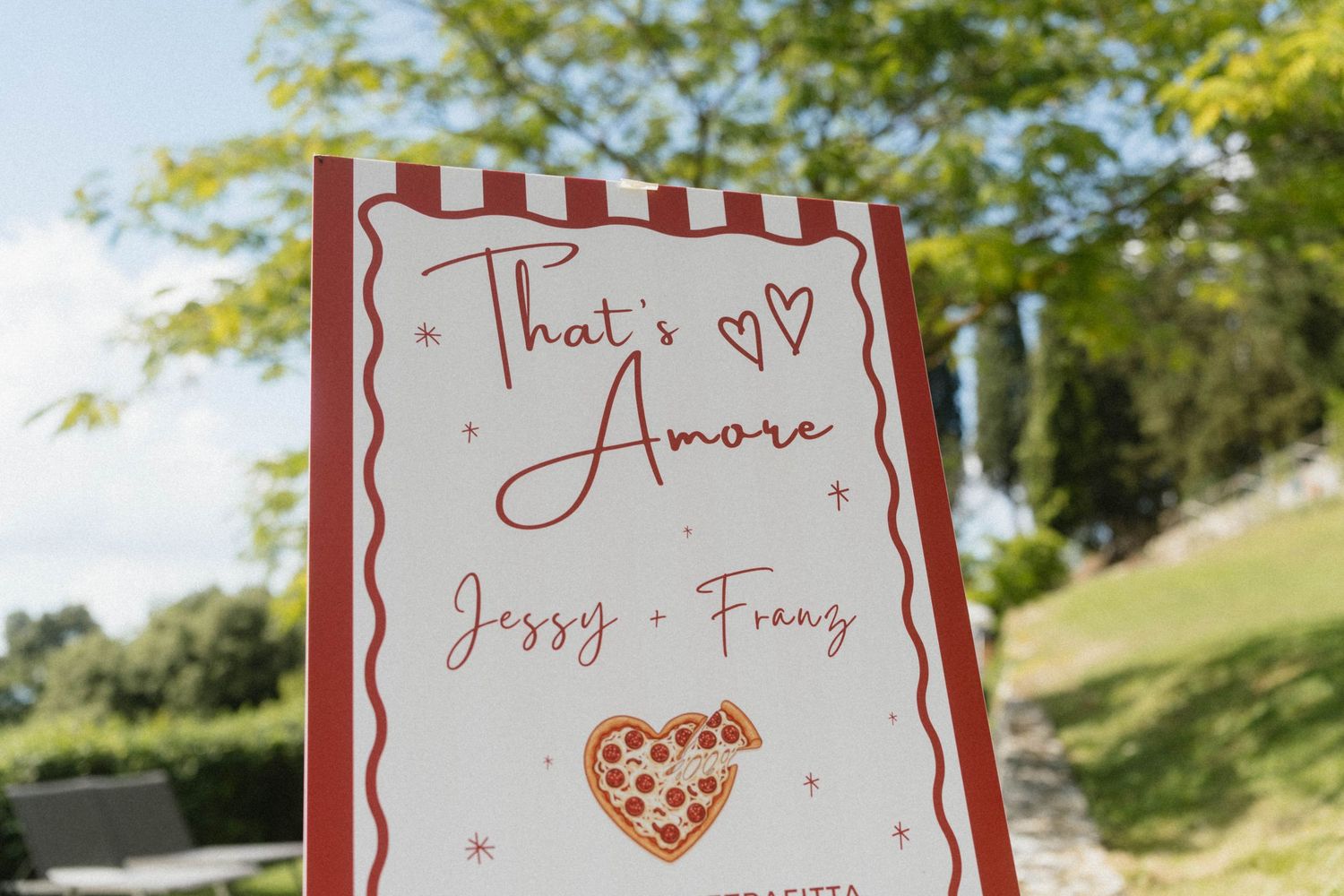 Wedding welcome sign reading That's Amore with Jessy and Frans names and heart-shaped pizza illustration outdoors.