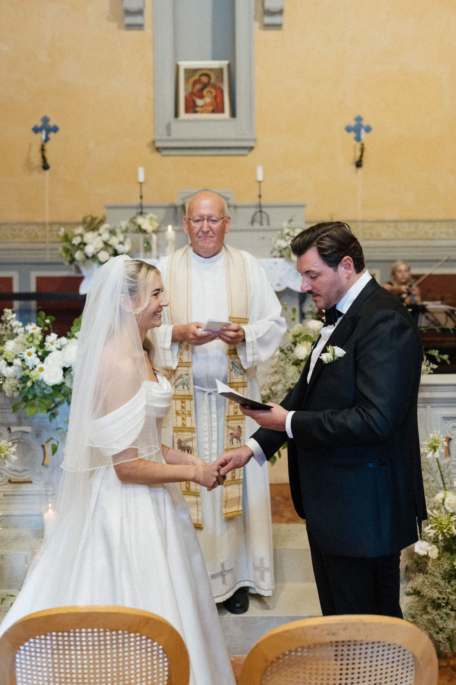 Bride and groom exchange vows at church altar with priest officiating, surrounded by white floral arrangements.