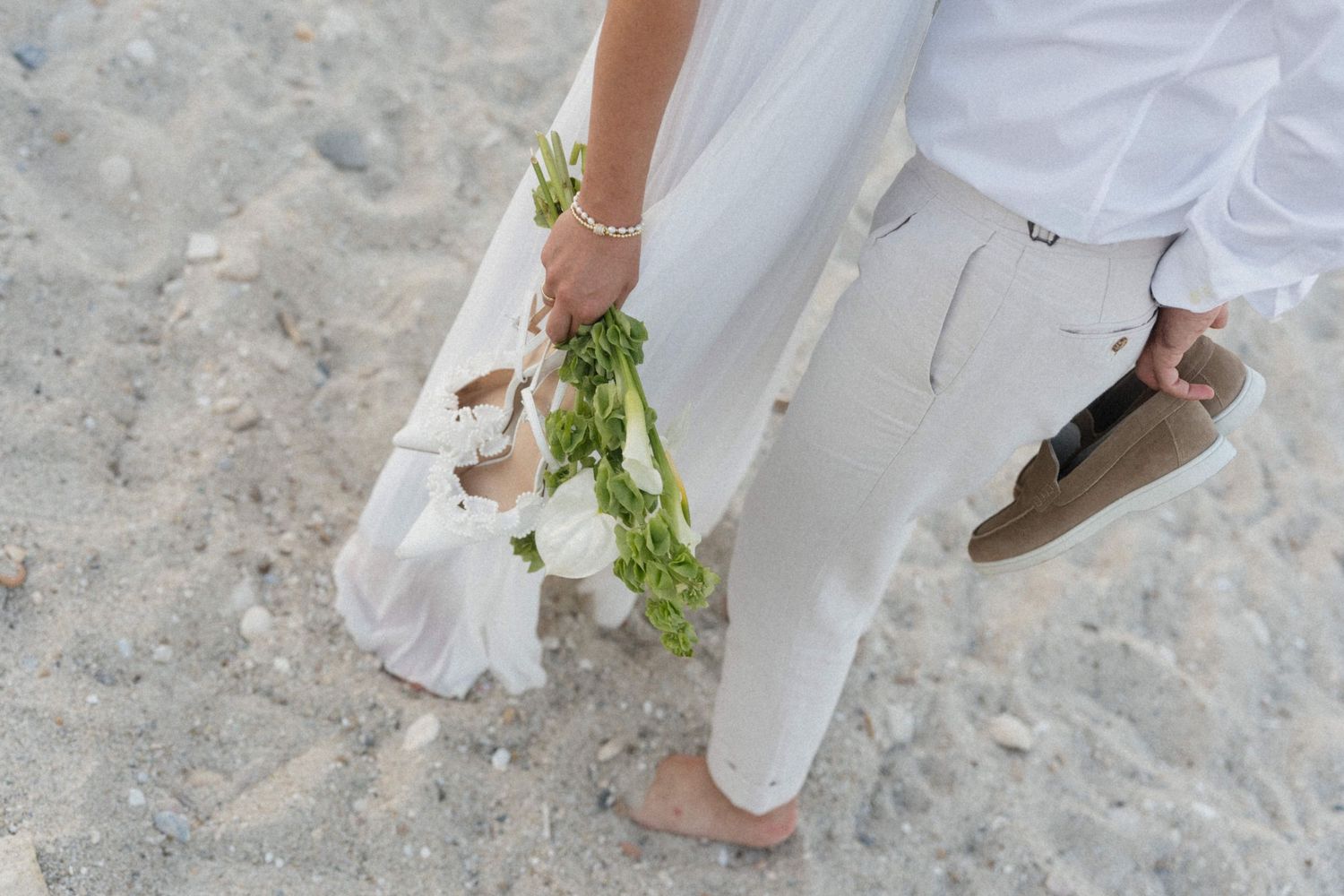 Barefoot couple in white wedding attire walking on sandy beach, bride holding white floral bouquet and shoes.