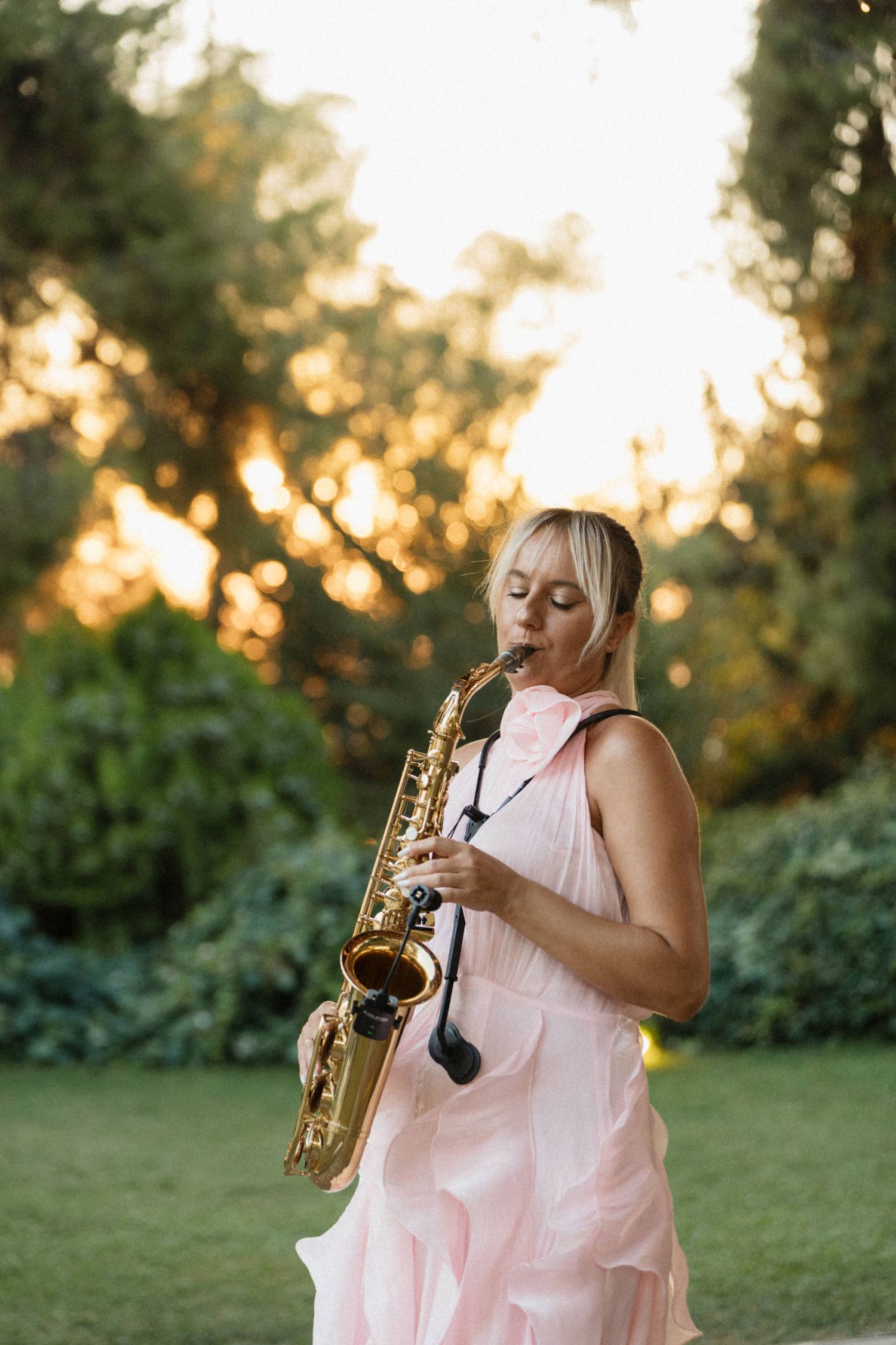 A blonde woman in a pink dress plays a golden saxophone outdoors in a lush green garden at sunset.