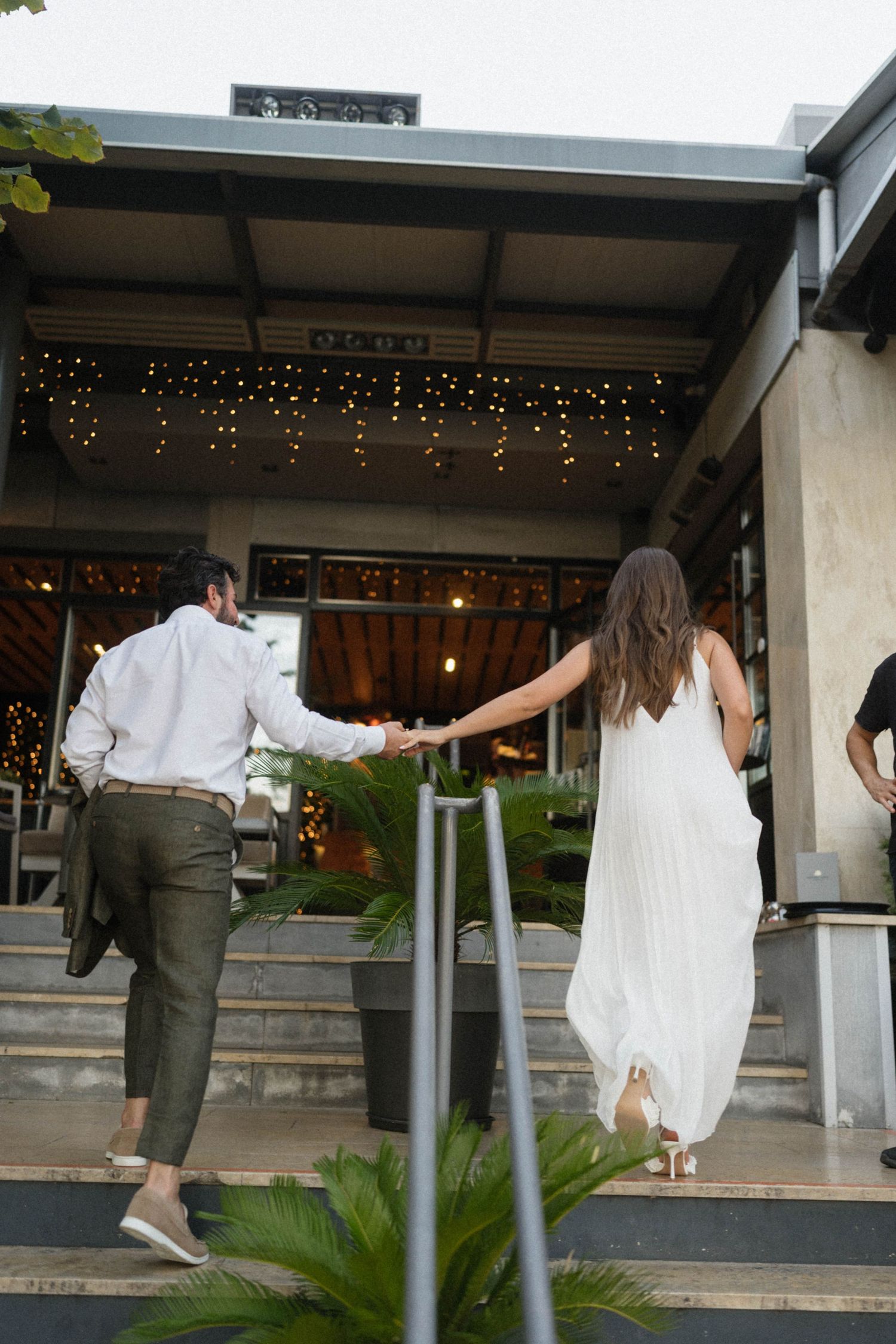 Couple holding hands walking up steps into a venue decorated with fairy lights, woman in white dress and man in green pants.