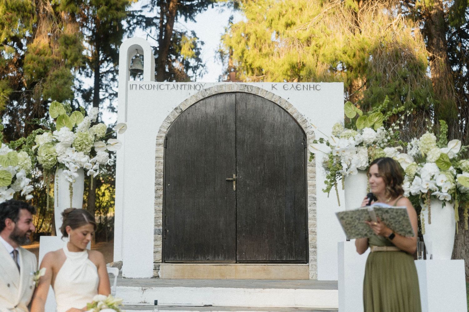 Outdoor wedding ceremony in front of a stone chapel with white floral arrangements and a celebrant reading vows.