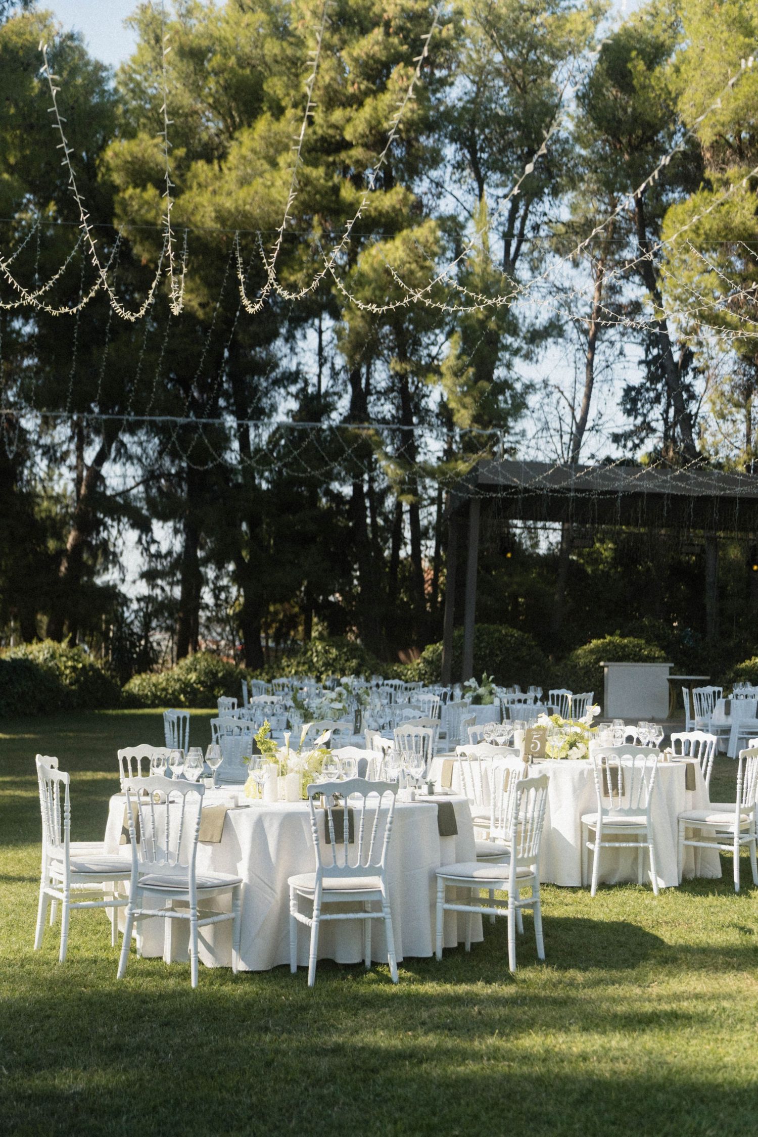 Elegant outdoor wedding reception with white round tables, chiavari chairs, and string lights among lush green trees.