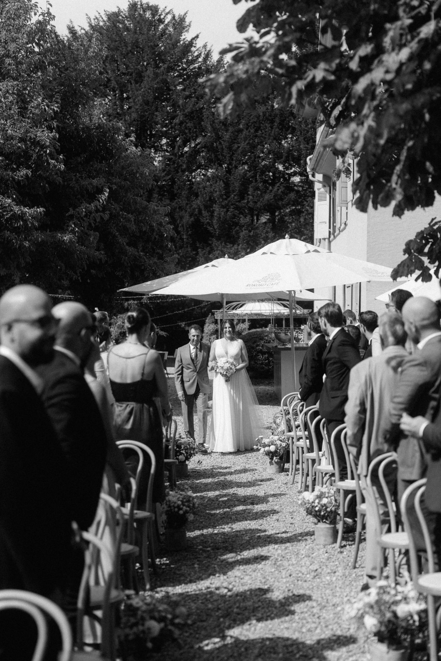 Black and white photo of bride walking down outdoor aisle lined with guests and chairs.