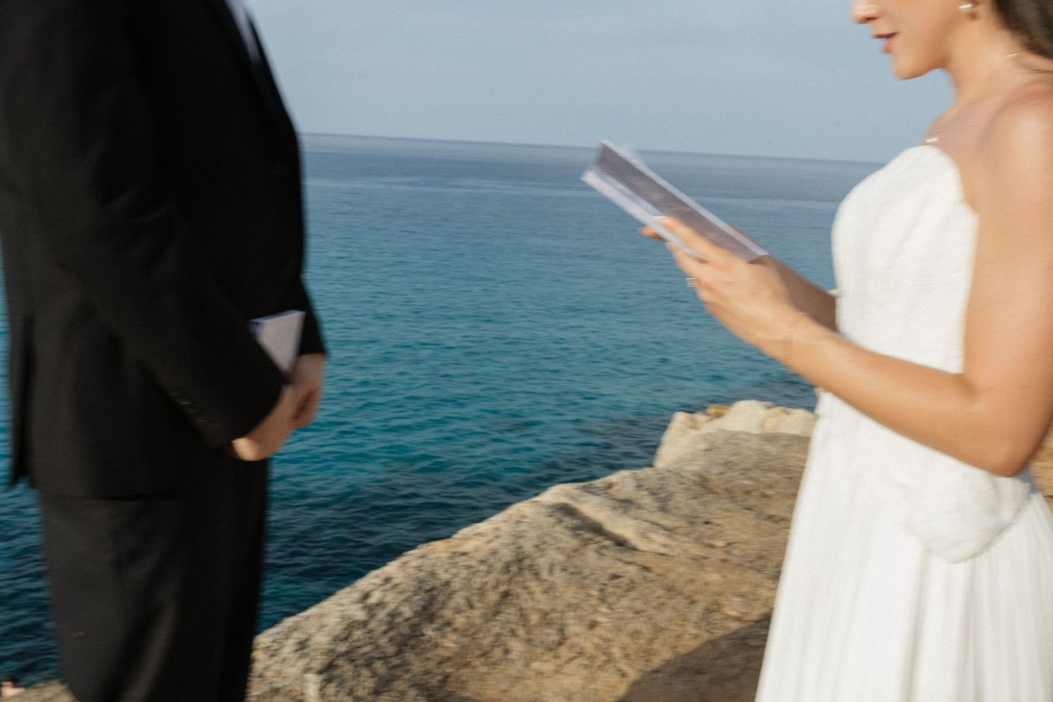 Bride in a white dress reads her vows from a booklet beside a groom in a black suit on a rocky cliff above the blue sea.