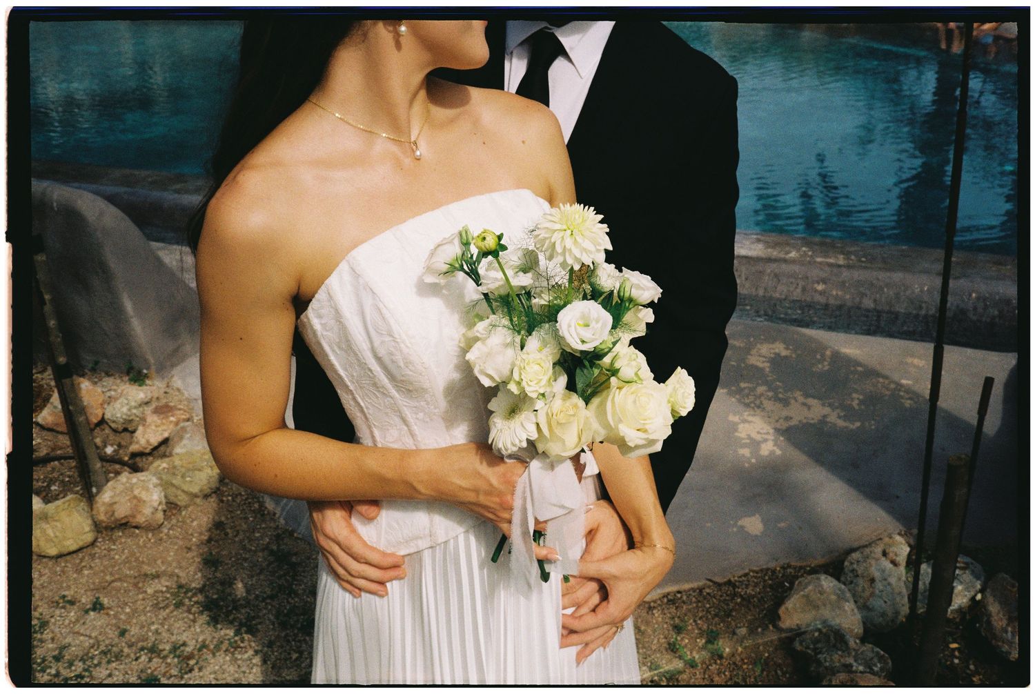 Bride in a white strapless gown holds a bouquet of white roses while embraced by a groom in a dark suit beside a sparkling pool.