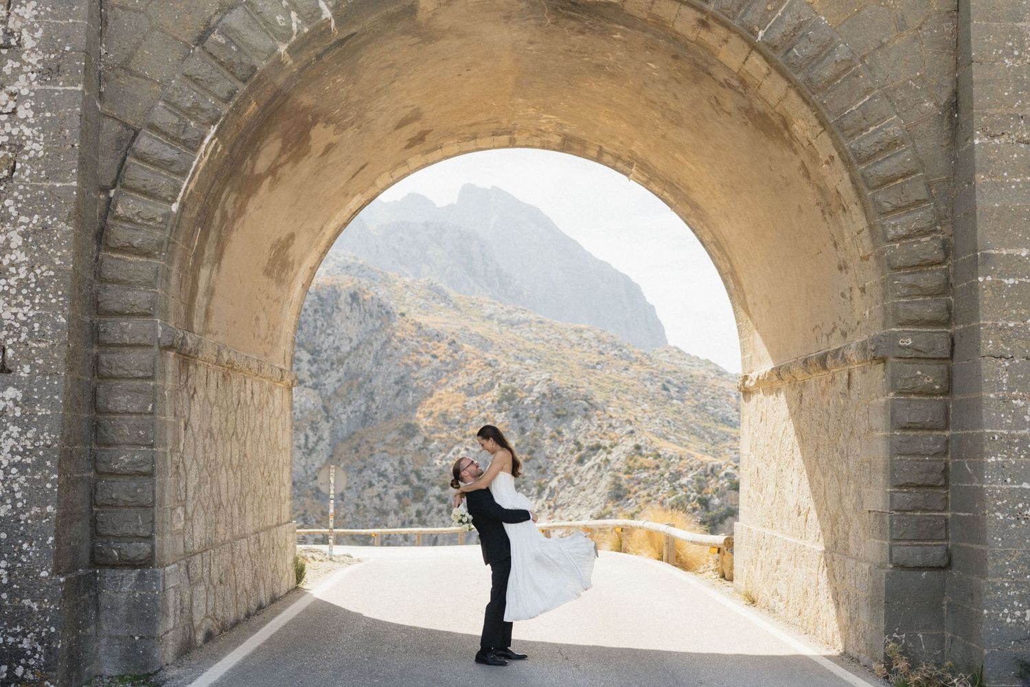 Bride in a white gown and groom in a dark suit embrace beneath a large stone tunnel arch with a rocky mountain view beyond.