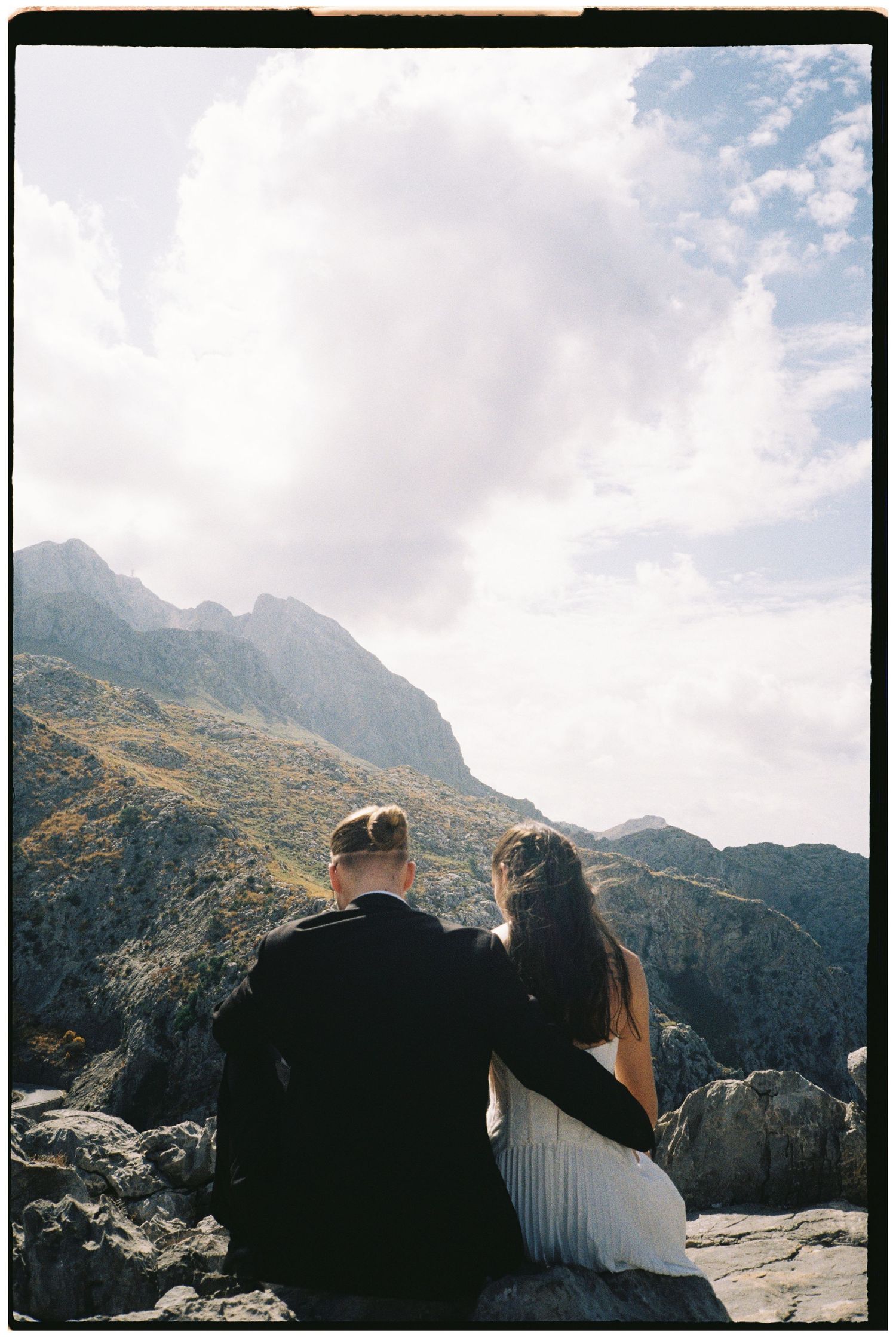 Couple in wedding attire sit close on a rocky mountain ledge, gazing at rugged peaks under a cloudy sky in a film-style photo.