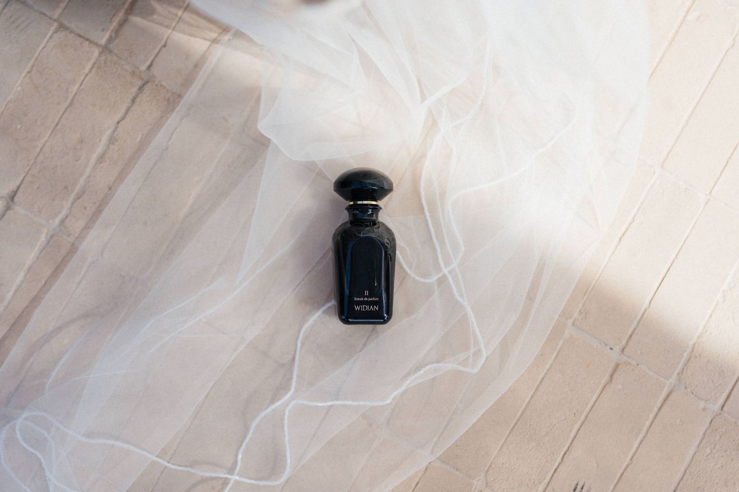 Black glass perfume bottle with a rounded cap resting on a sheer white bridal veil over a light weathered wooden floor.