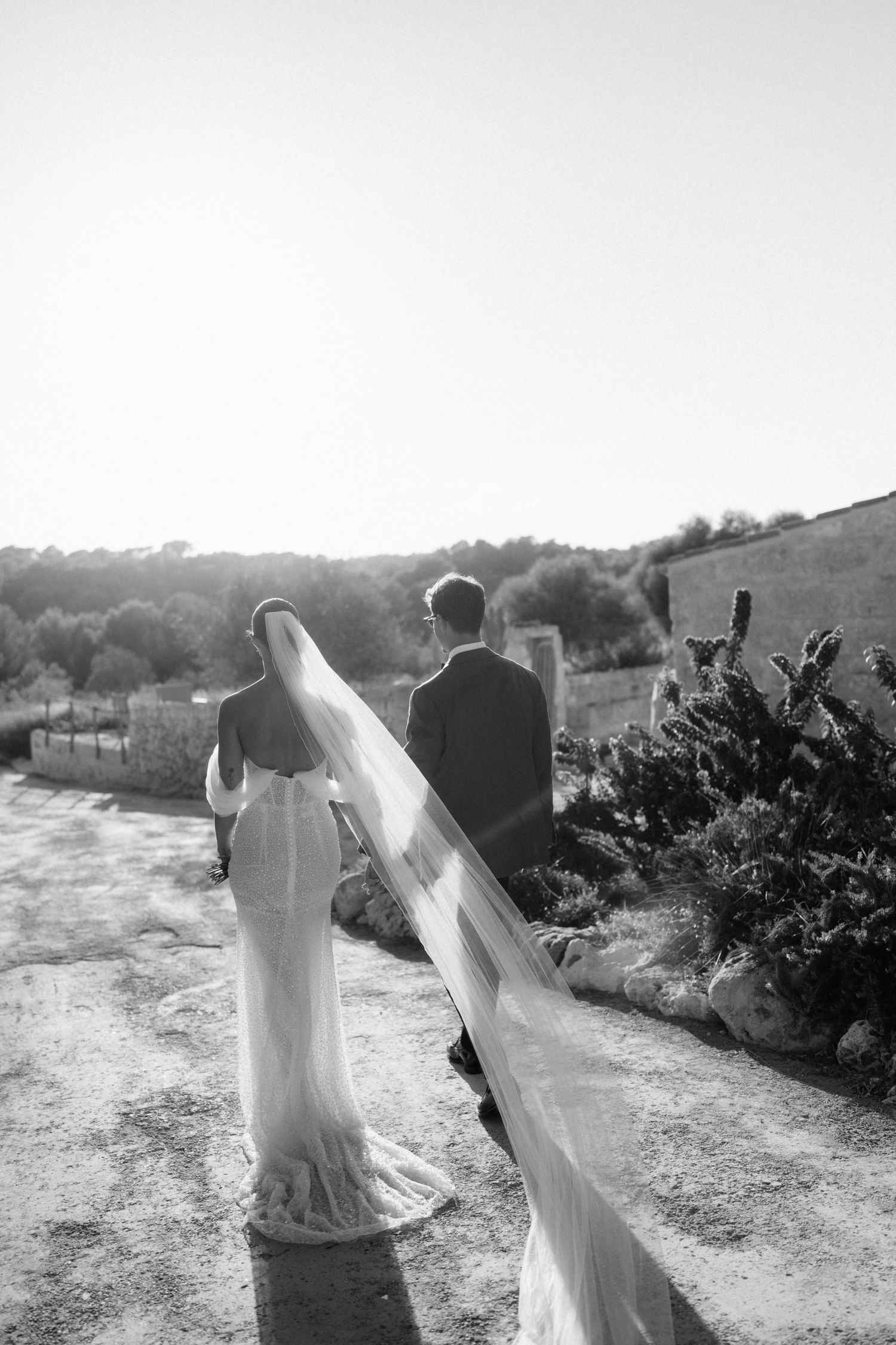 Black and white photo of a bride in a long lace-trimmed veil walking with her groom along a rustic path.