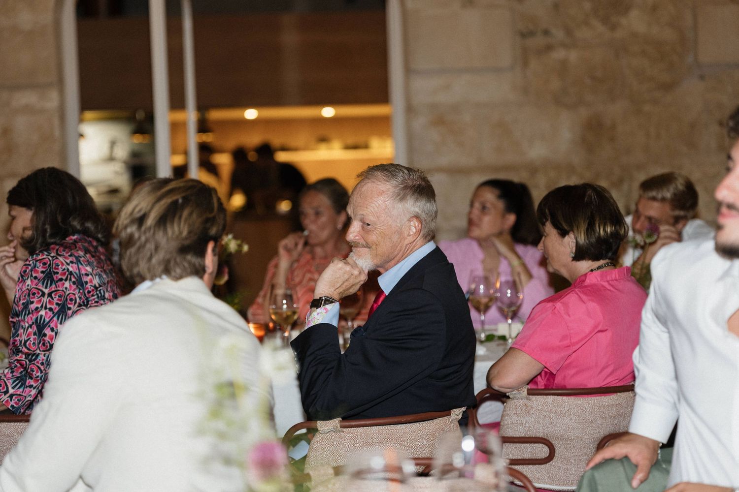 Smiling wedding guests in colorful outfits enjoy conversation at a reception table, an older man in a dark suit in focus.
