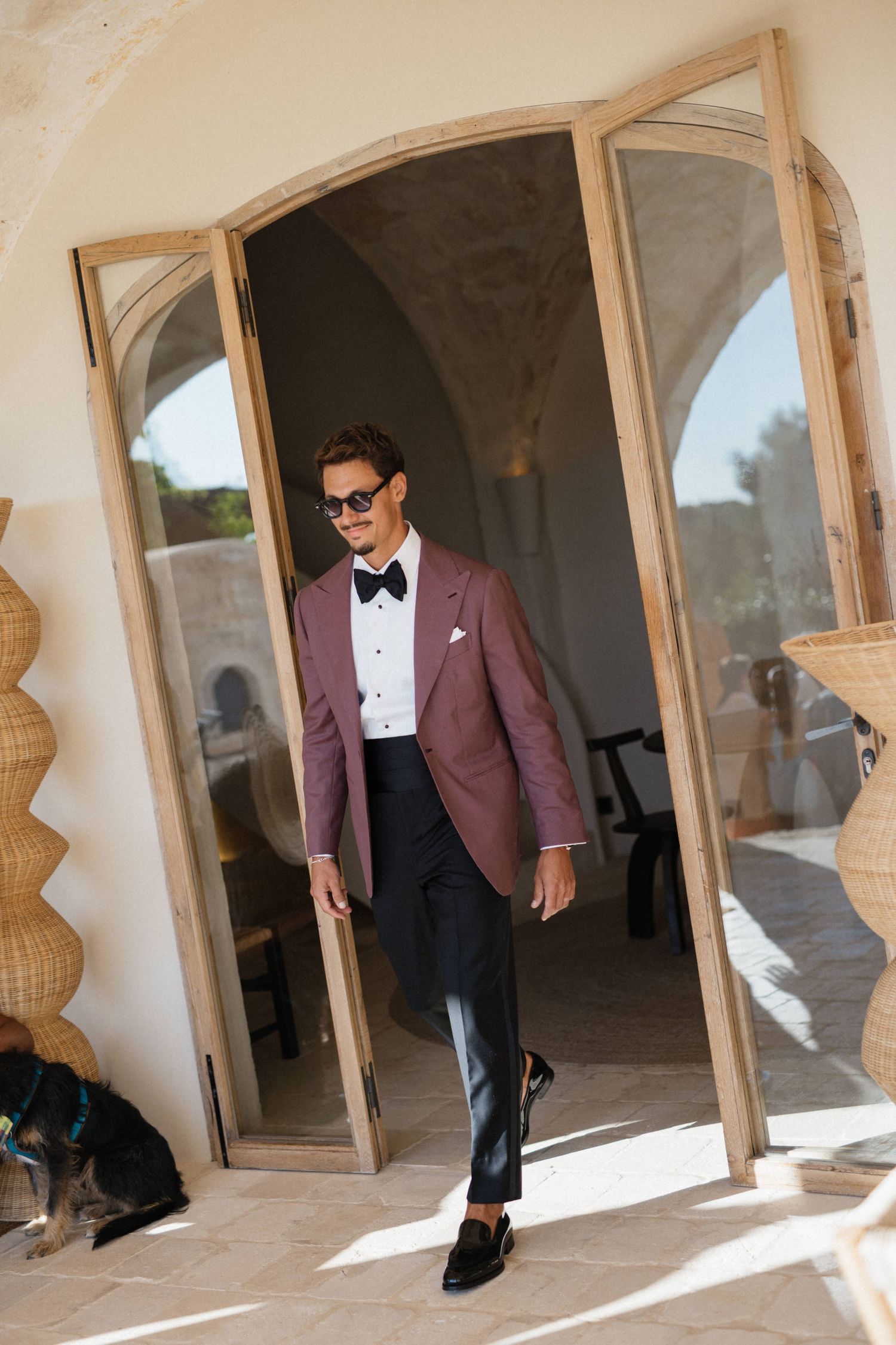 Stylish groom in a burgundy tuxedo jacket, bow tie and sunglasses steps through open arched doors onto a sunny terrace.