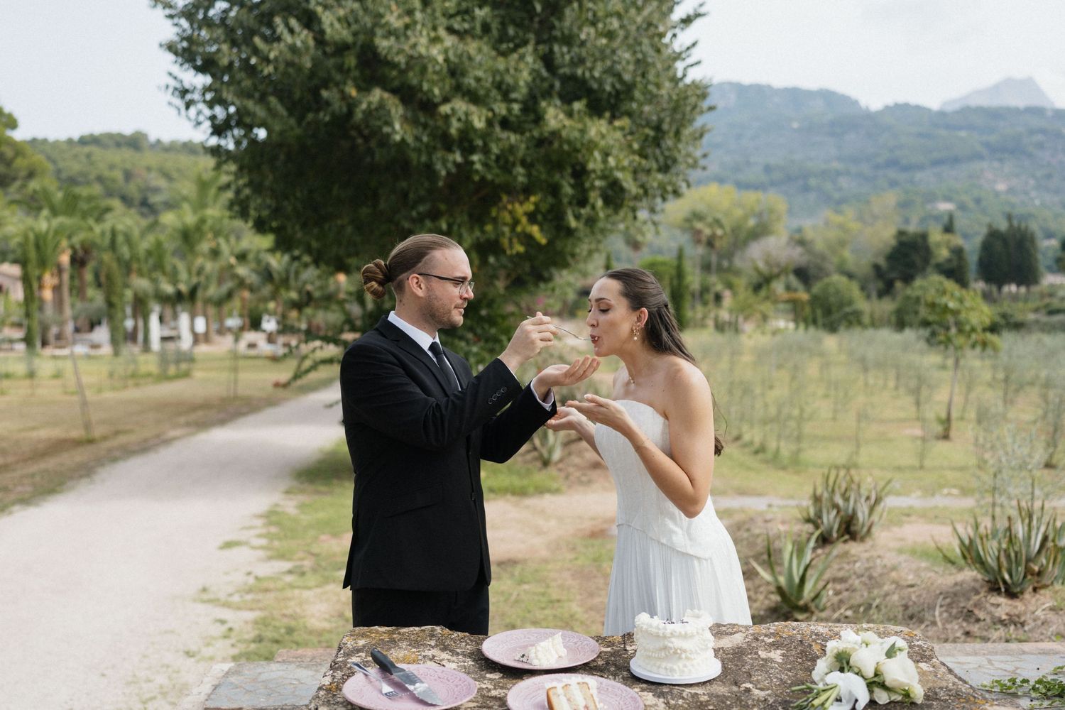 Bride in white dress and groom in dark suit feed each other wedding cake at an outdoor table overlooking a scenic vineyard and mountains.