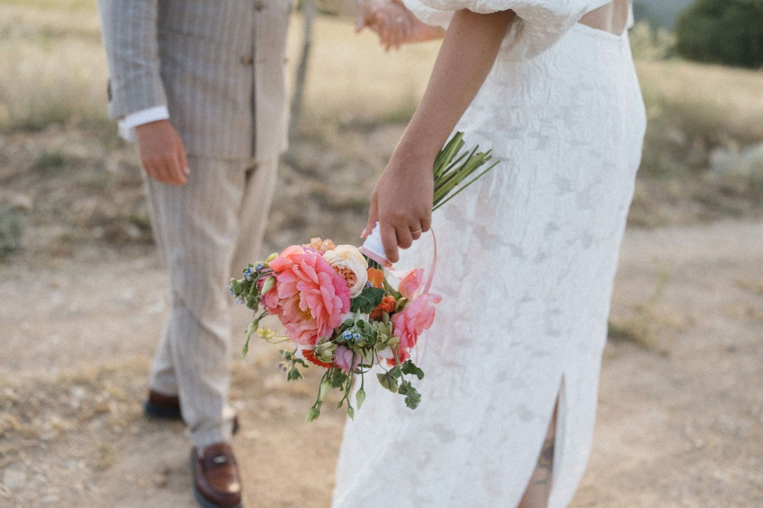 Bride in lace gown holding pink tropical bouquet walks with groom in tan suit on dry field path.
