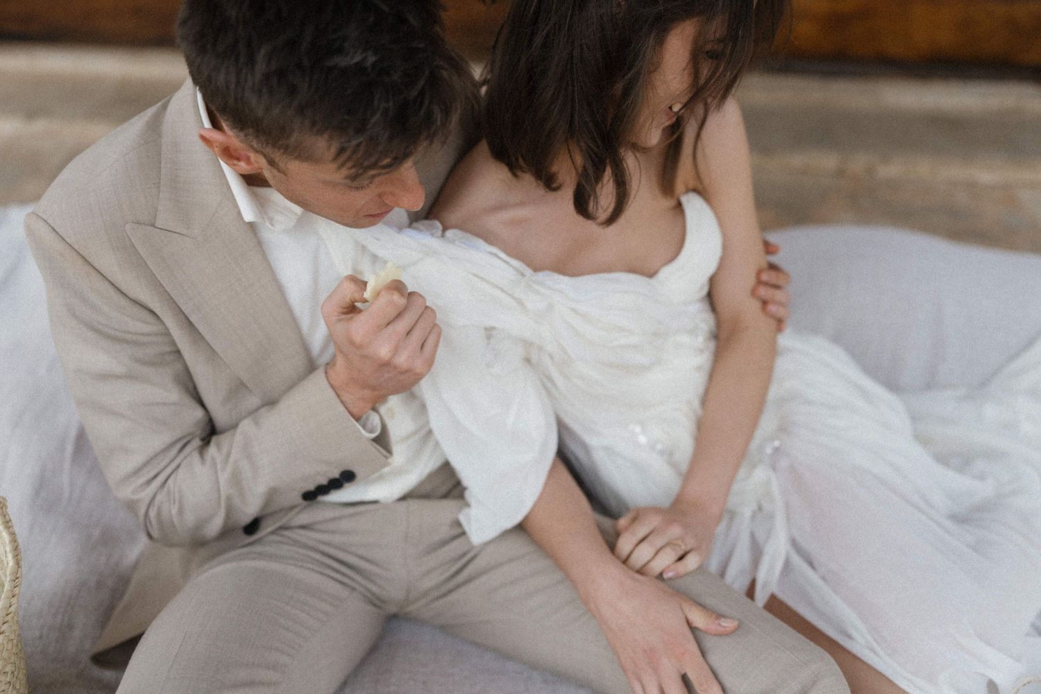 Groom in beige suit kisses bride's hand while she sits in flowing white wedding dress.
