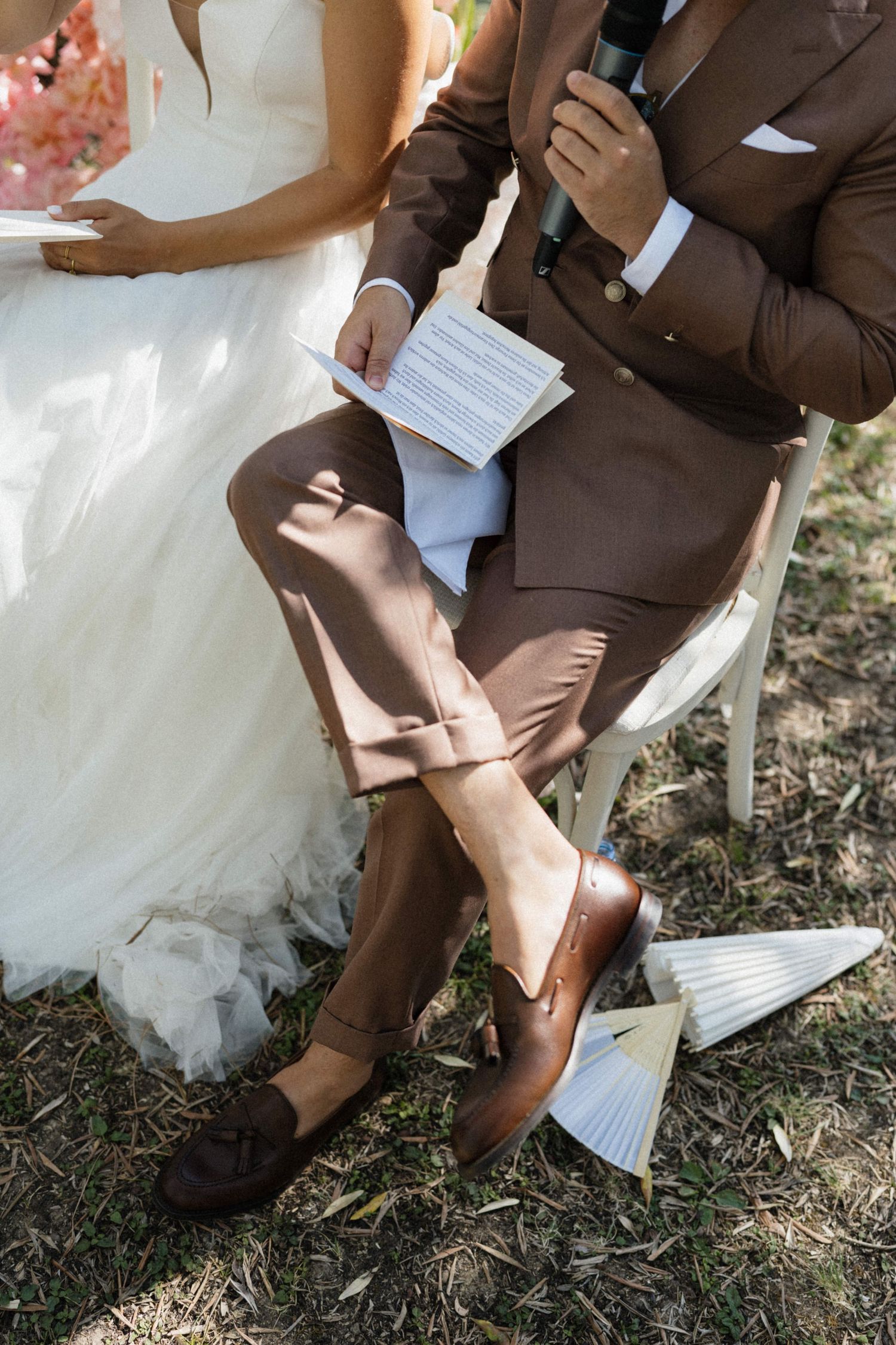 Groom in brown suit holding vows paper at outdoor wedding ceremony, bride in white dress seated beside him.