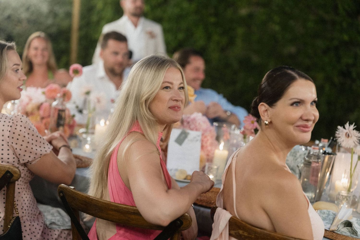 Women in pink and blue dresses smile and enjoy conversation at an outdoor wedding reception with candles.