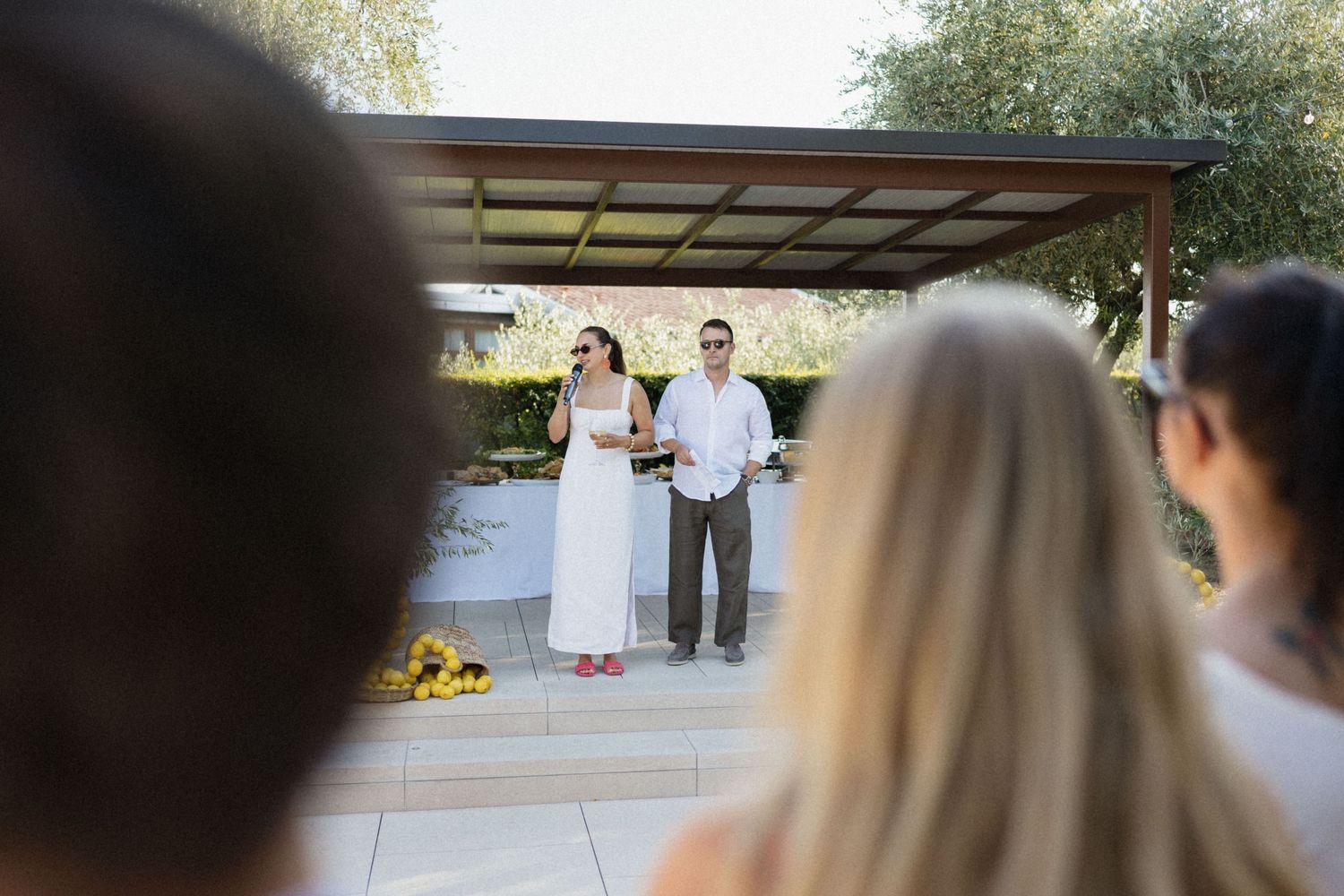 Couple in white attire standing under pergola during outdoor wedding ceremony with guests watching.