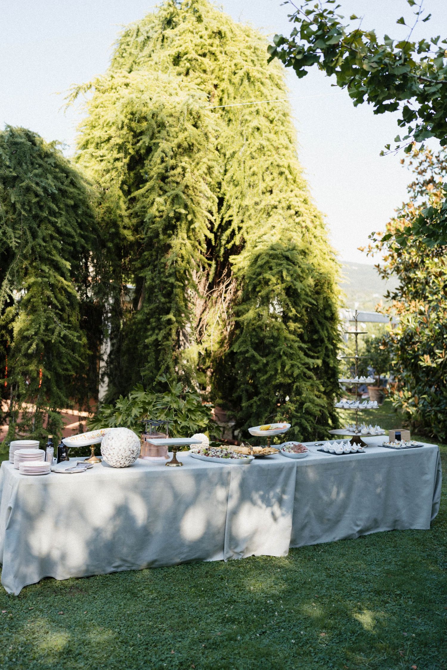 Elegant outdoor buffet table with gray linen set in a lush garden with tall cypress trees.