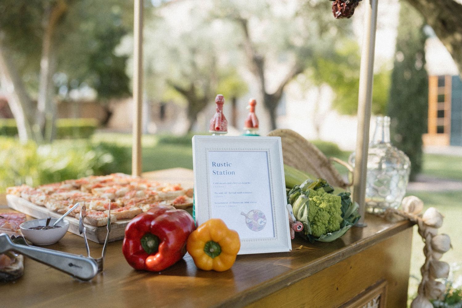 Outdoor wedding food display table with colorful bell peppers, fresh vegetables, pizza, and a framed menu sign.